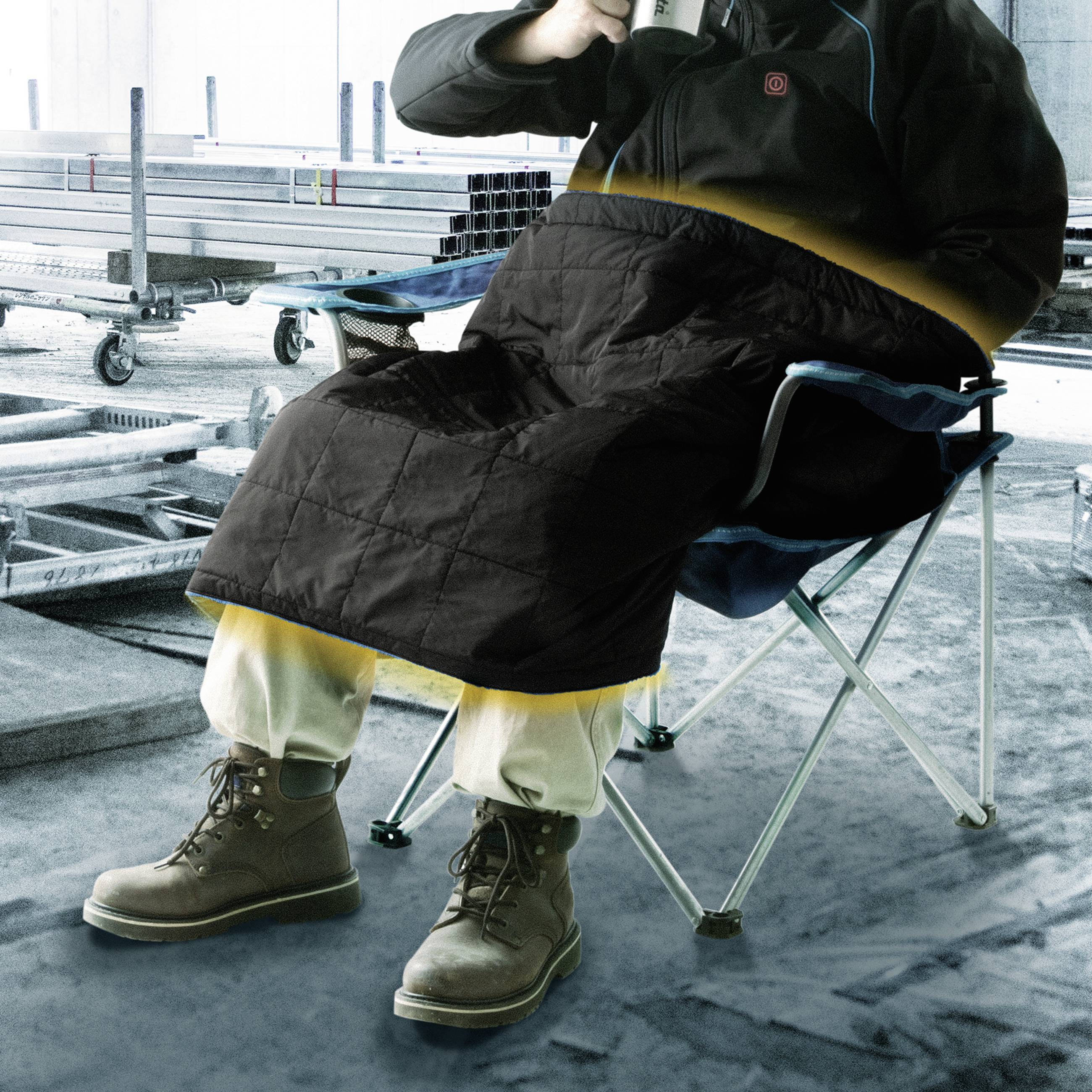Eine Person sitzt auf einem Klappstuhl in einem industriellen Lager, trägt eine isolierte Decke und trinkt aus einem Becher.