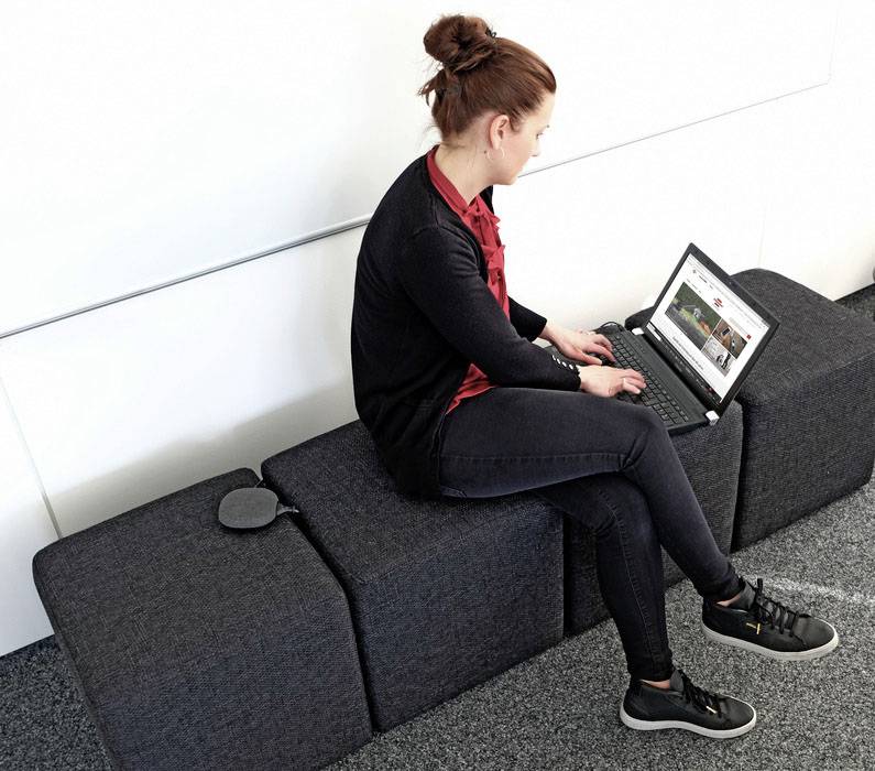 Eine Frau sitzt auf einem grauen Sofa und arbeitet an einem Laptop, der auf ihren Knien liegt. Im Hintergrund eine weiße Wand.