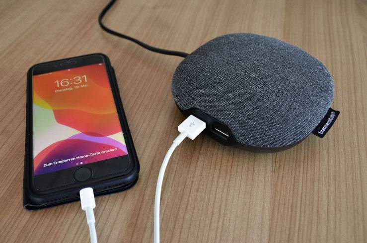 Ein Smartphone mit Ladebildschirm ist über ein USB-Kabel mit einem tragbaren Lautsprecher verbunden, der auf einem Holztisch liegt.