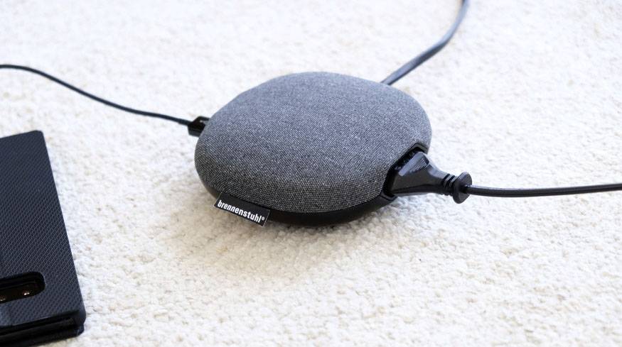 Ein graues, rundes Ladegerät mit textilbezogenem Design auf einem hellen Teppich. Zwei Kabel sind angesteckt. Ein schwarzes Objekt liegt daneben.