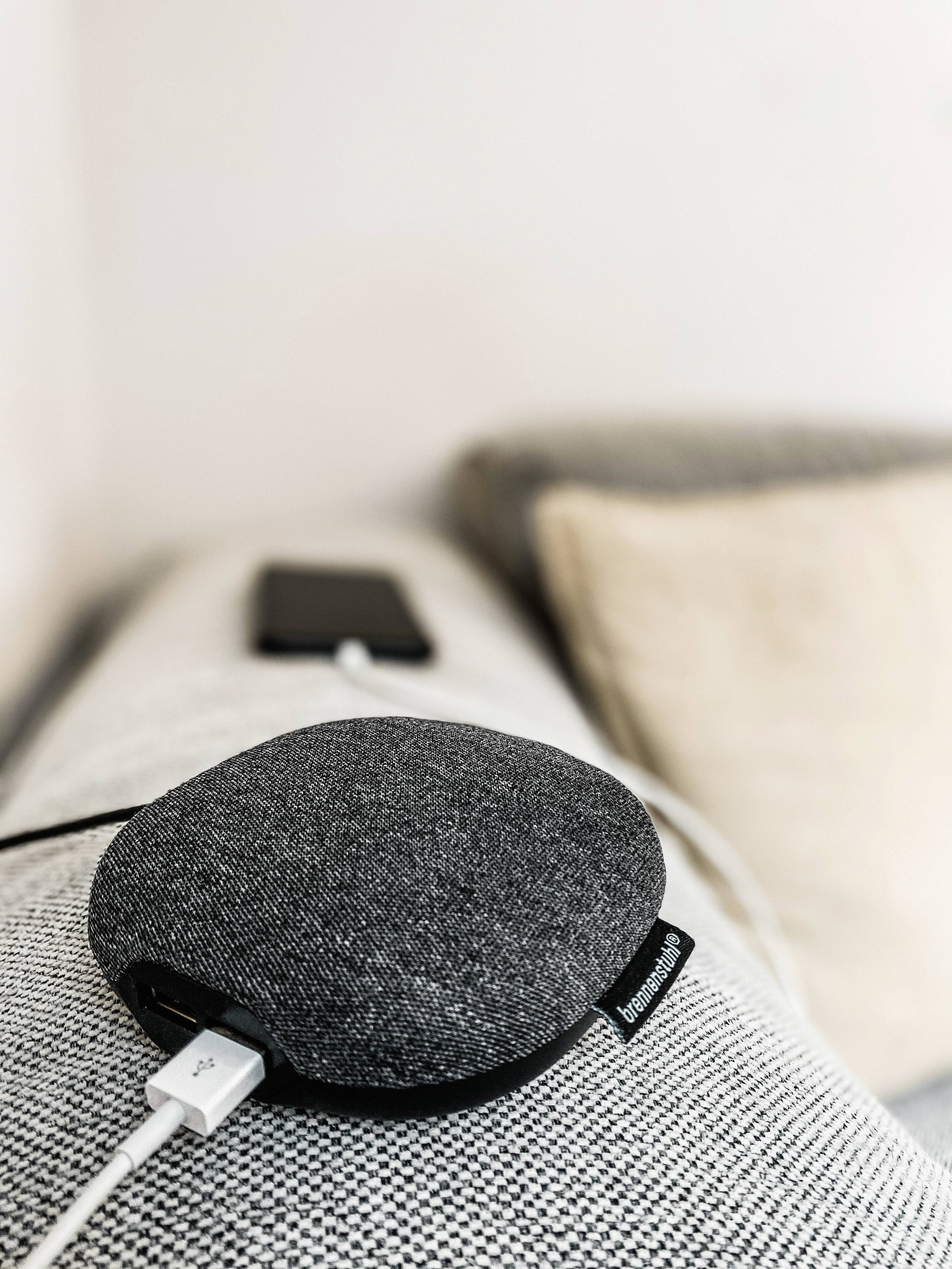 Ein grauer, runder, tragbarer Lautsprecher liegt auf einer hellen Couch. Im Hintergrund ist ein unscharfes Smartphone erkennbar.