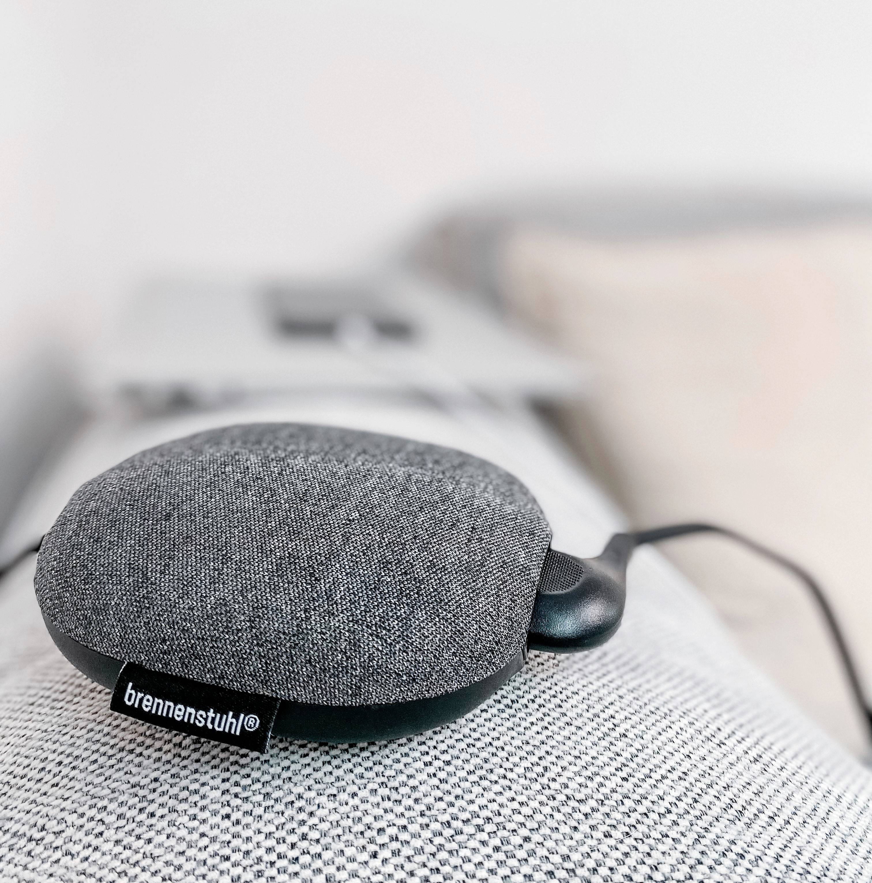 Ein grauer, ovaler Bluetooth-Lautsprecher liegt auf einem Stoffsofa. Das Lautsprecher-Etikett zeigt die Marke 'brennenstuhl'.
