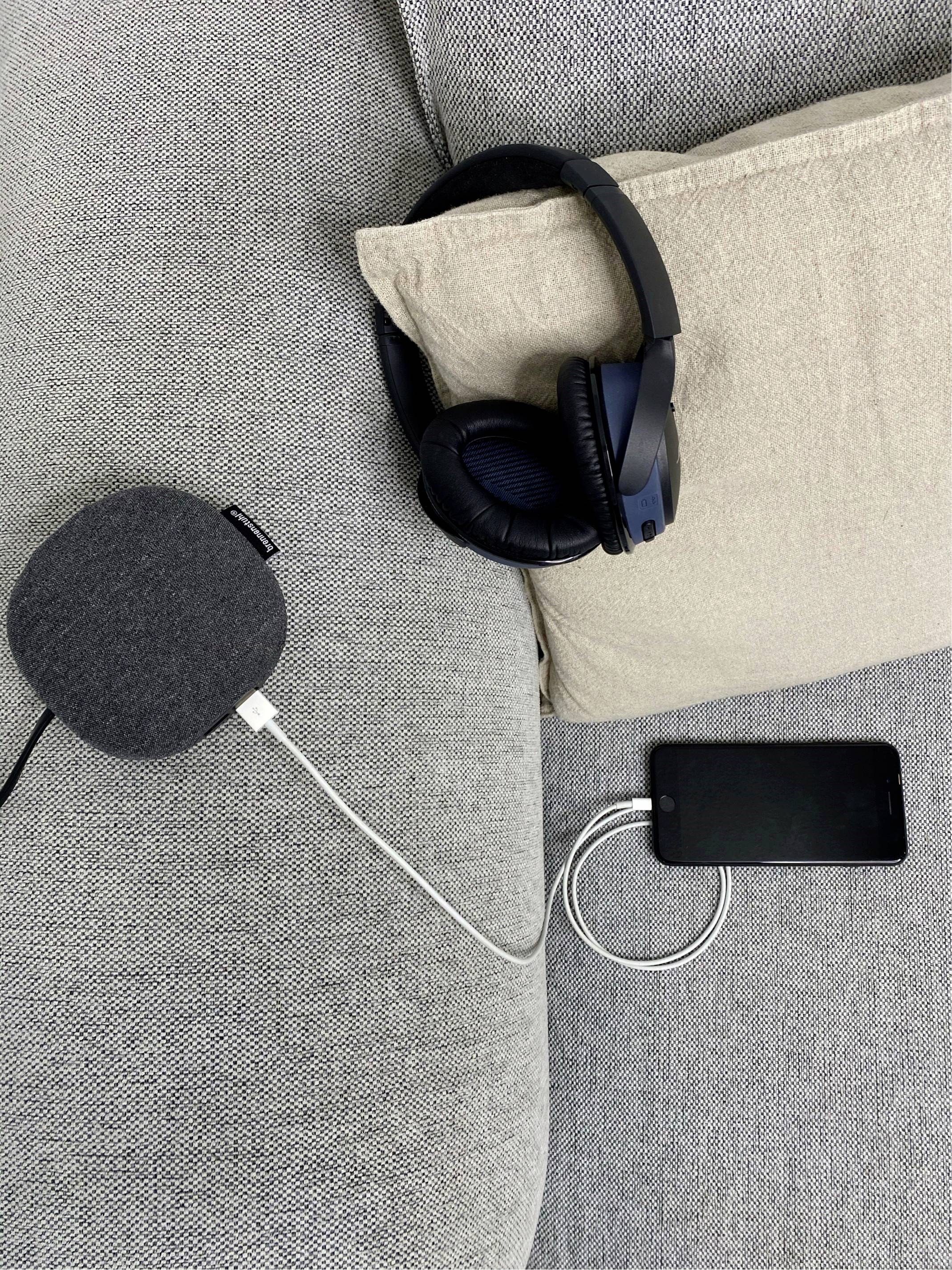 Ein Smartphone, ein Ladegerät und Kopfhörer liegen auf einem grauen Sofa, verbunden mit einem Ladegerät, das an einen Lautsprecher angeschlossen ist.
