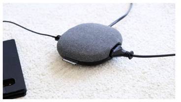 Ein grauer, kugelförmiger Lautsprecher mit zwei verbundenen Kabeln liegt auf einem hellen Teppich. Daneben liegt ein schwarzes Smartphone.