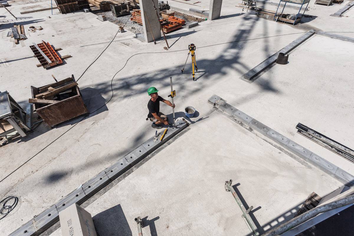 Arbeiter mit grünem Helm misst auf einer Baustelle mit einem Vermessungsgerät. Schatten eines Krans auf Betonboden.