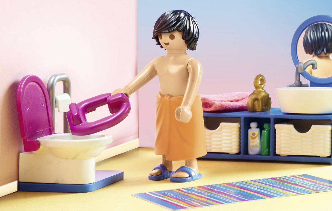 Eine Playmobil-Figur in einem Badezimmer hält einen Toilettendeckel. Sie trägt ein orangefarbenes Handtuch und steht neben einem Waschbecken.