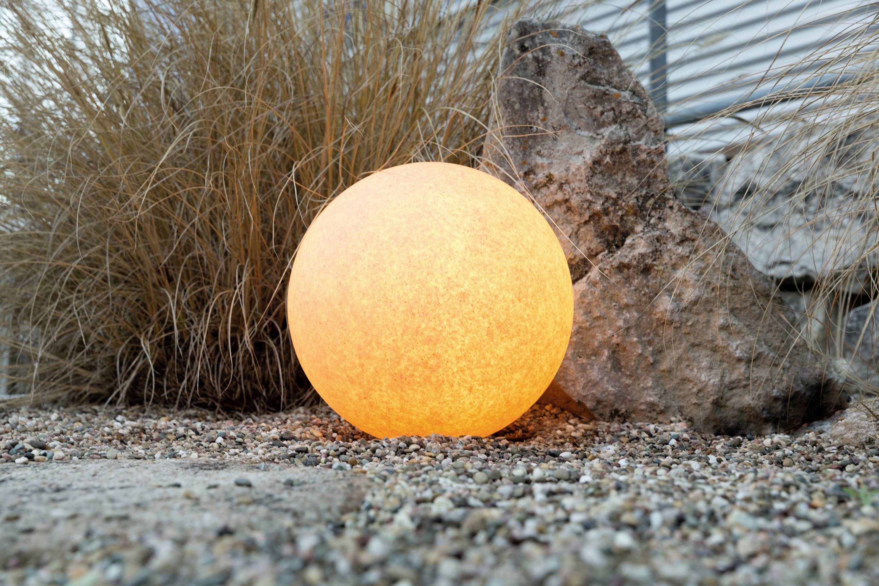 Leuchtende Kugellampe im Garten, umgeben von Steinen und Gräsern, erzeugt warmes Licht und stimmungsvolle Abendatmosphäre.