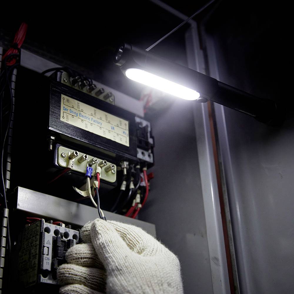 Eine Person mit Handschuhen arbeitet an einer elektrischen Schalttafel unter einer starken Leuchte in einem dunklen Raum.