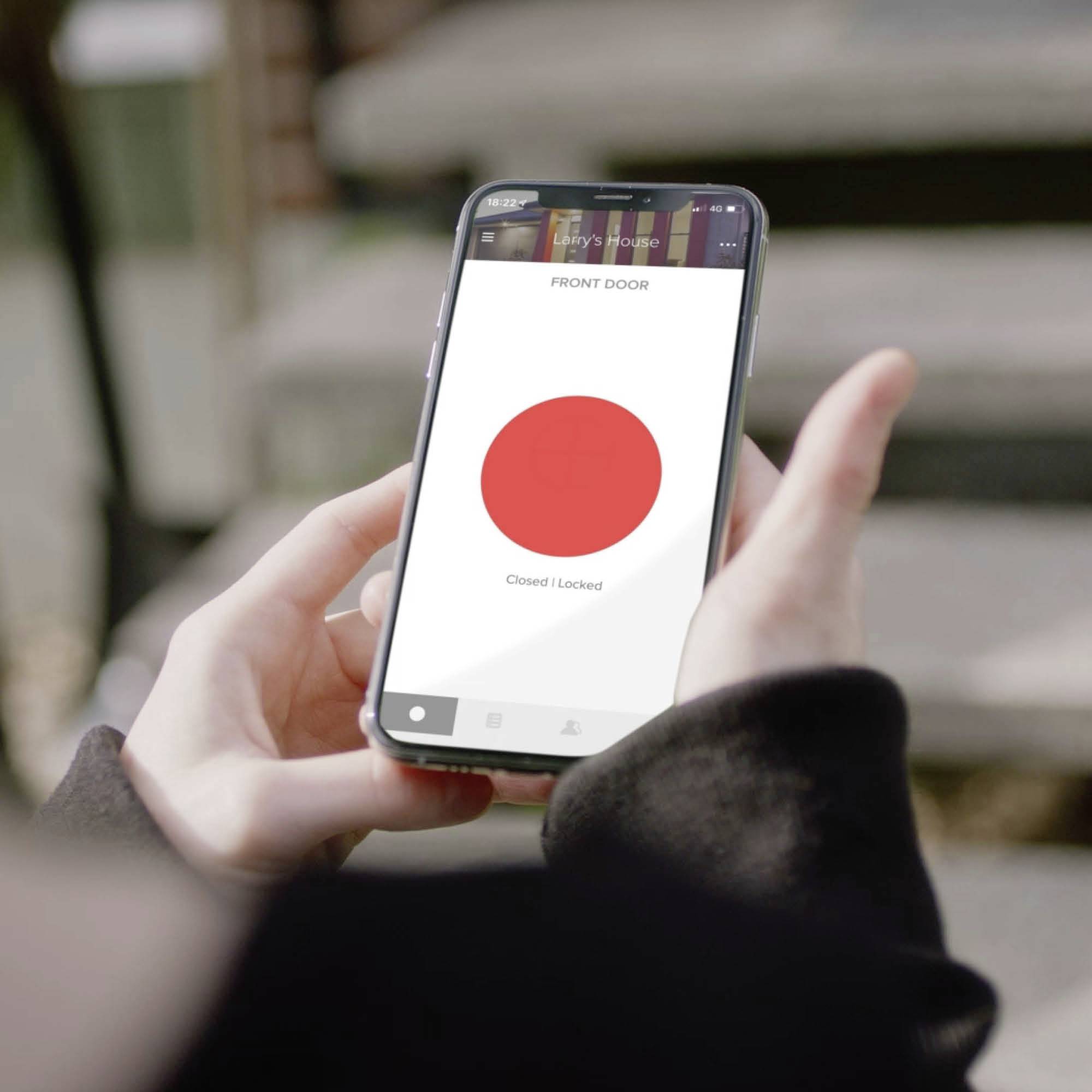 Person hält ein Smartphone mit einer App, die anzeigt: 'Front Door - Closed/Locked', roter Punkt in der Mitte. Anastasia im Hintergrund.