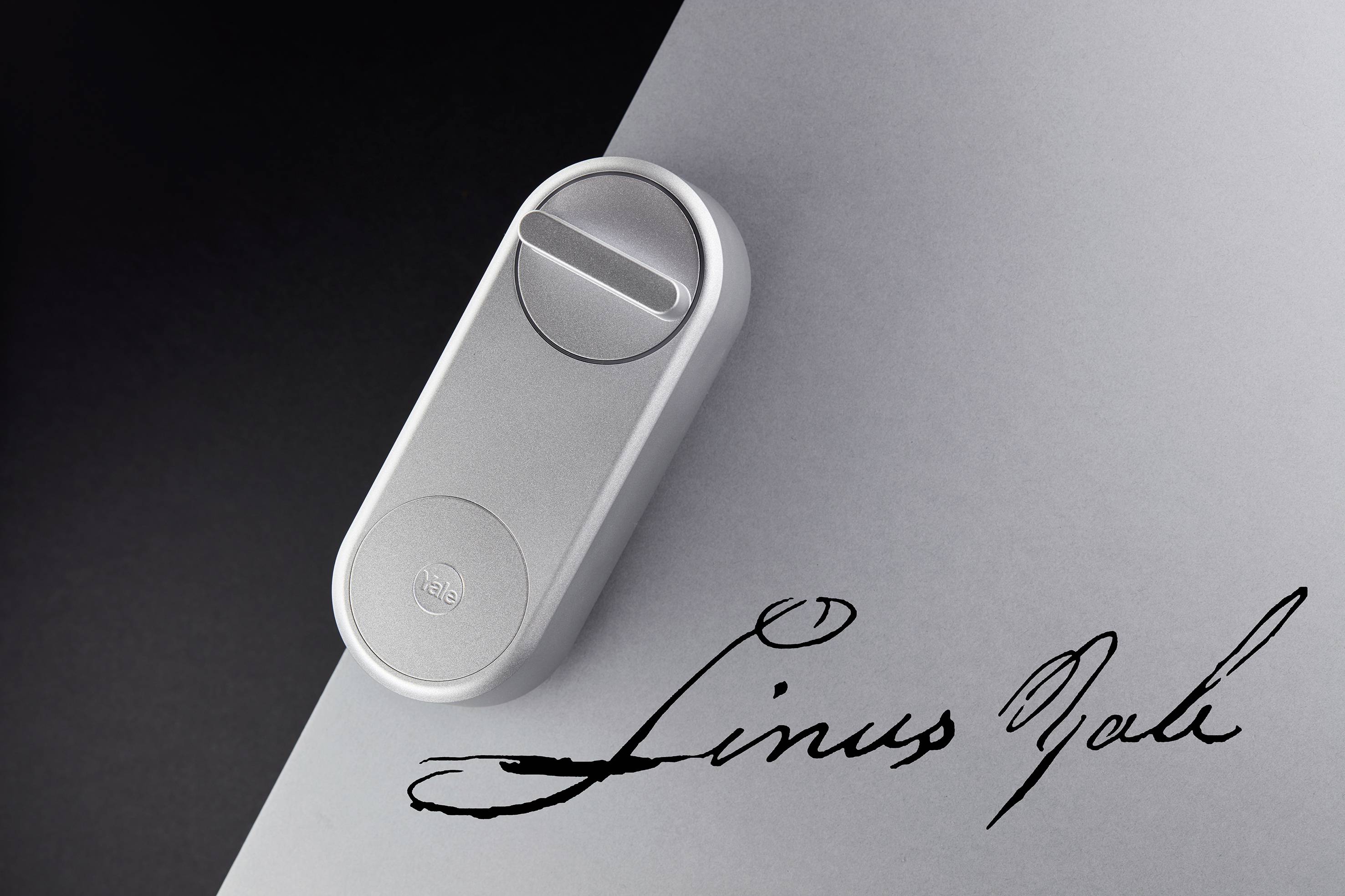 Ein silbernes Smart-Türschloss auf grauem Hintergrund. Daneben ist eine stilisierte Unterschrift in schwarzer Schreibschrift zu sehen.