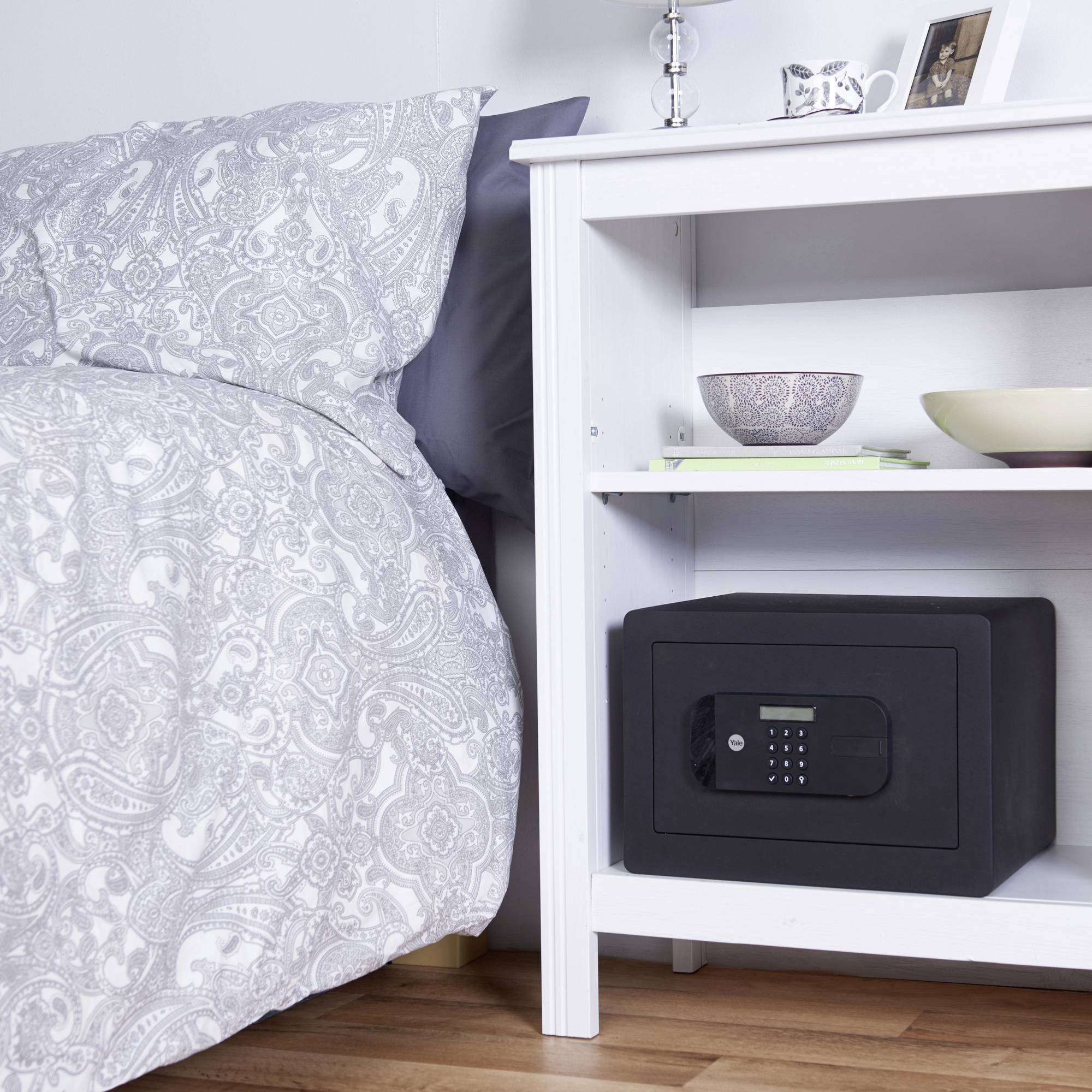 Ein kleiner Safe steht in einem weißen Nachttischregal neben einem Bett mit grauer gemusterter Bettwäsche.