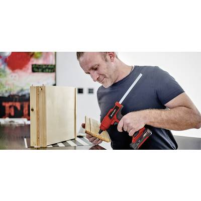 Ein Mann befestigt ein Holzbrett mit einem Akku-Bohrschrauber an einer Box. Im Hintergrund ist unscharf ein buntes Bild sichtbar.