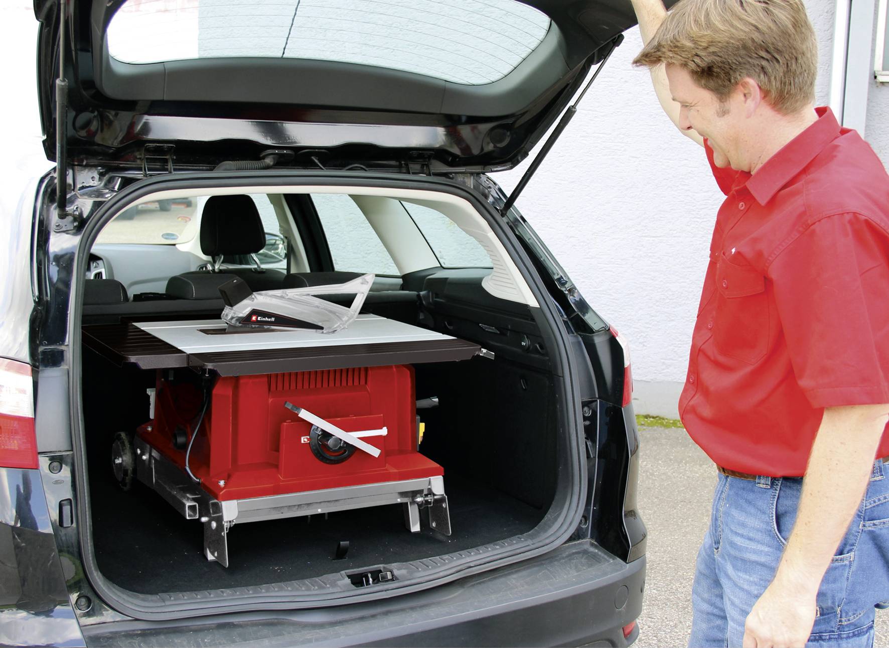 Ein Mann steht neben einem Auto mit geöffneter Kofferraumklappe, in dem eine rote Tischsäge transportiert wird.