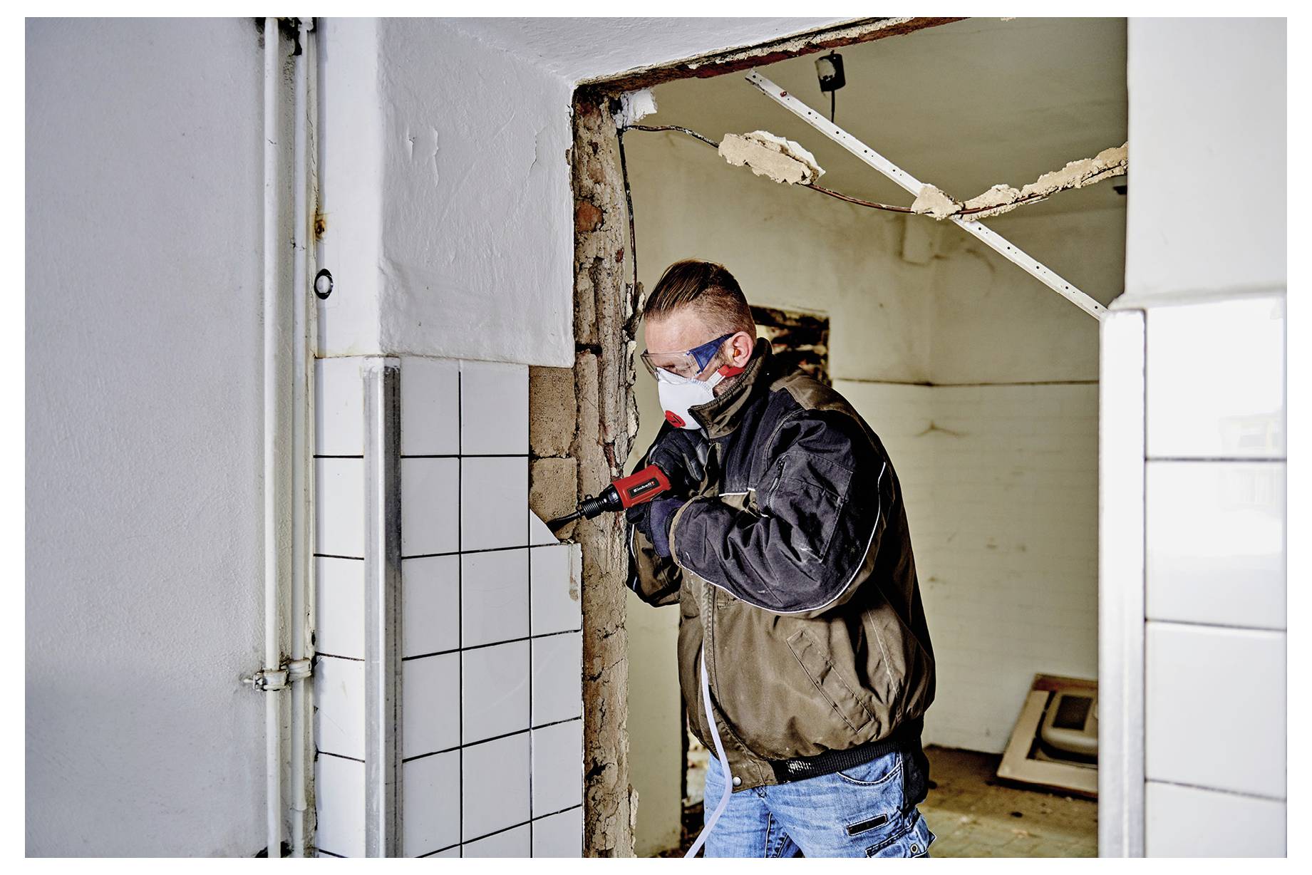 Arbeiter mit Maske und Handschuhen benutzt einen Elektroschrauber, um Fliesen aus einer Türöffnung während der Innenraumdemolition zu entfernen.