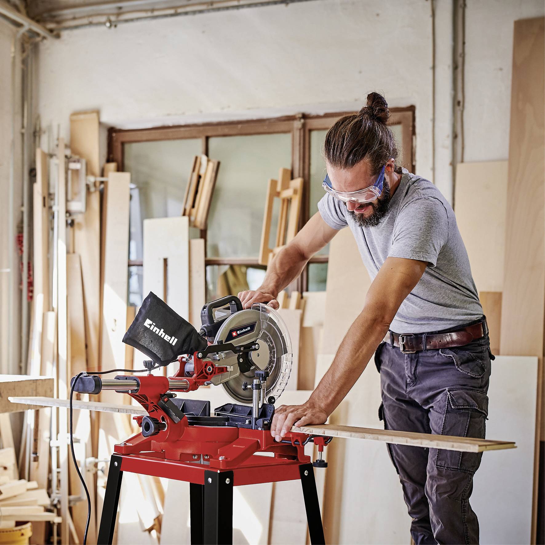 Eine Person in einer Werkstatt schneidet mit einer Kappsäge Holz. Im Hintergrund sind Holzplanken und Fensterrahmen zu sehen.