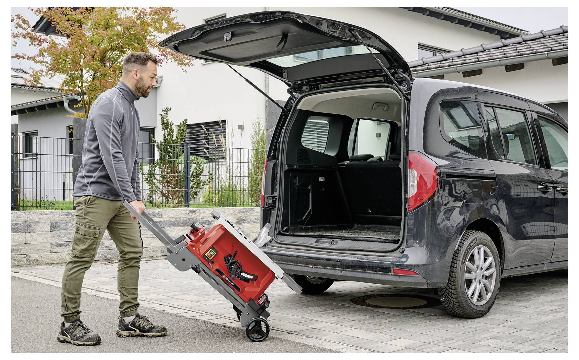 Eine Person in lockerer Kleidung lädt eine rote Maschine auf Rädern in den Kofferraum eines grauen Lieferwagens, der auf einer Einfahrt in einem Wohngebiet geparkt ist.