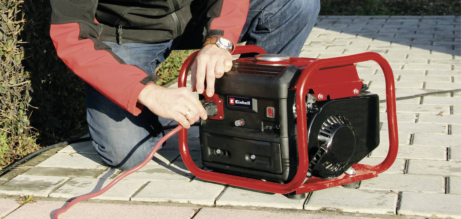 Eine Person verbindet ein Kabel mit einem roten tragbaren Generator auf einer gepflasterten Fläche im Freien.