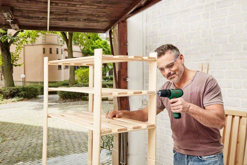 Ein Mann mit Schutzbrille benutzt eine Bohrmaschine, um ein Holzregal in einer offenen Werkstatt zusammenzubauen.