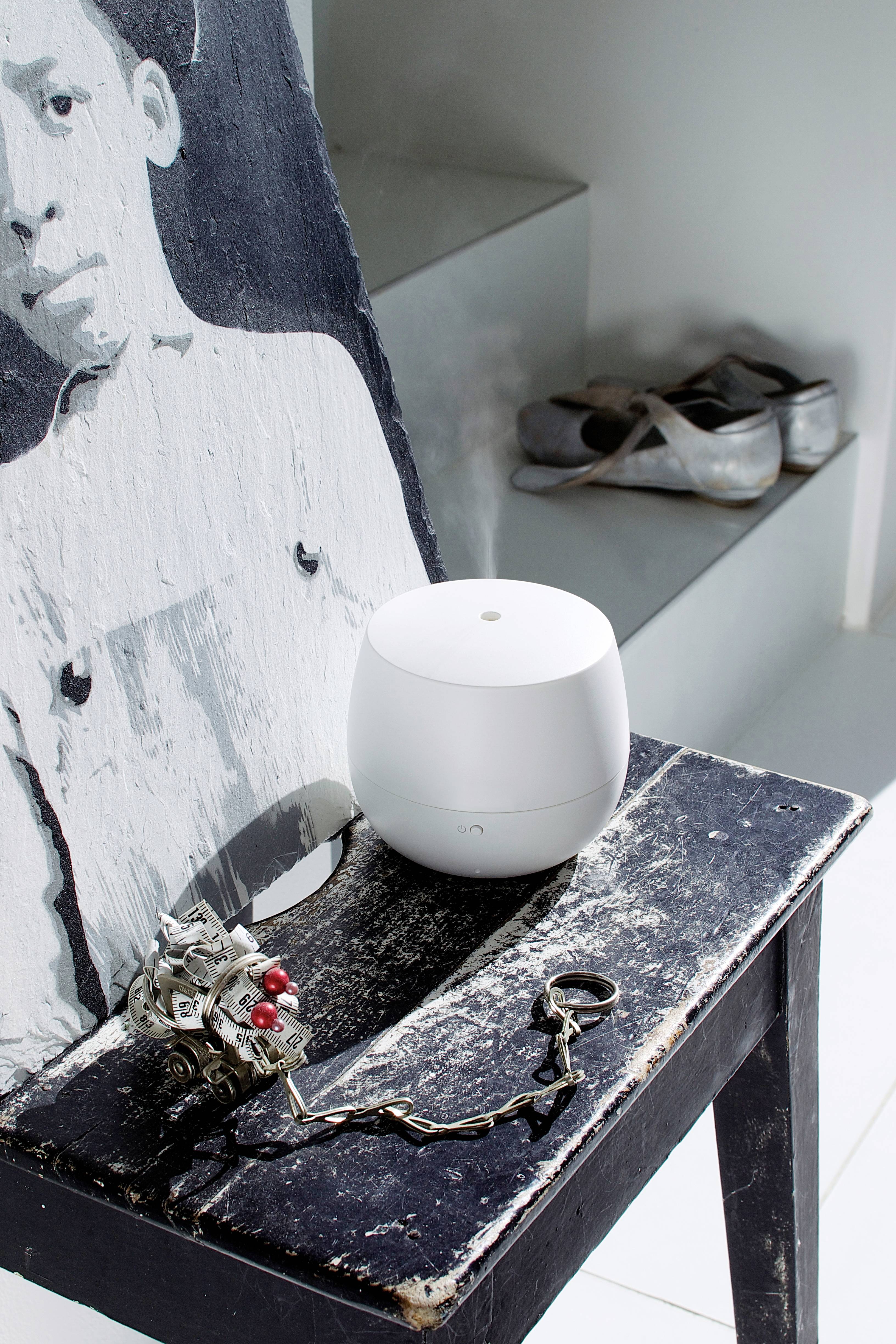 Ein weißer Lautsprecher steht auf einem alten Hocker neben einem kunstvollen Bild und einem mechanischen Gerät, im Hintergrund silberne Schuhe.