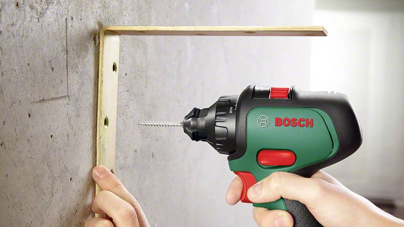 Eine Person montiert ein L-förmiges Metallwinkel an eine Wand mit einem grünen Akkuschrauber.