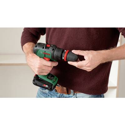 Eine Person hält eine kabellose Bohrmaschine und befestigt einen Bohreraufsatz. Sie trägt einen weinroten Pullover und eine Jeans.