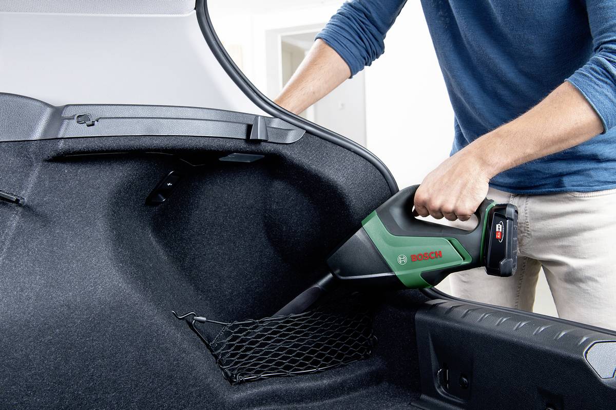 Person benutzt einen kleinen, grünen Handstaubsauger, um den Kofferraum eines Autos zu reinigen.