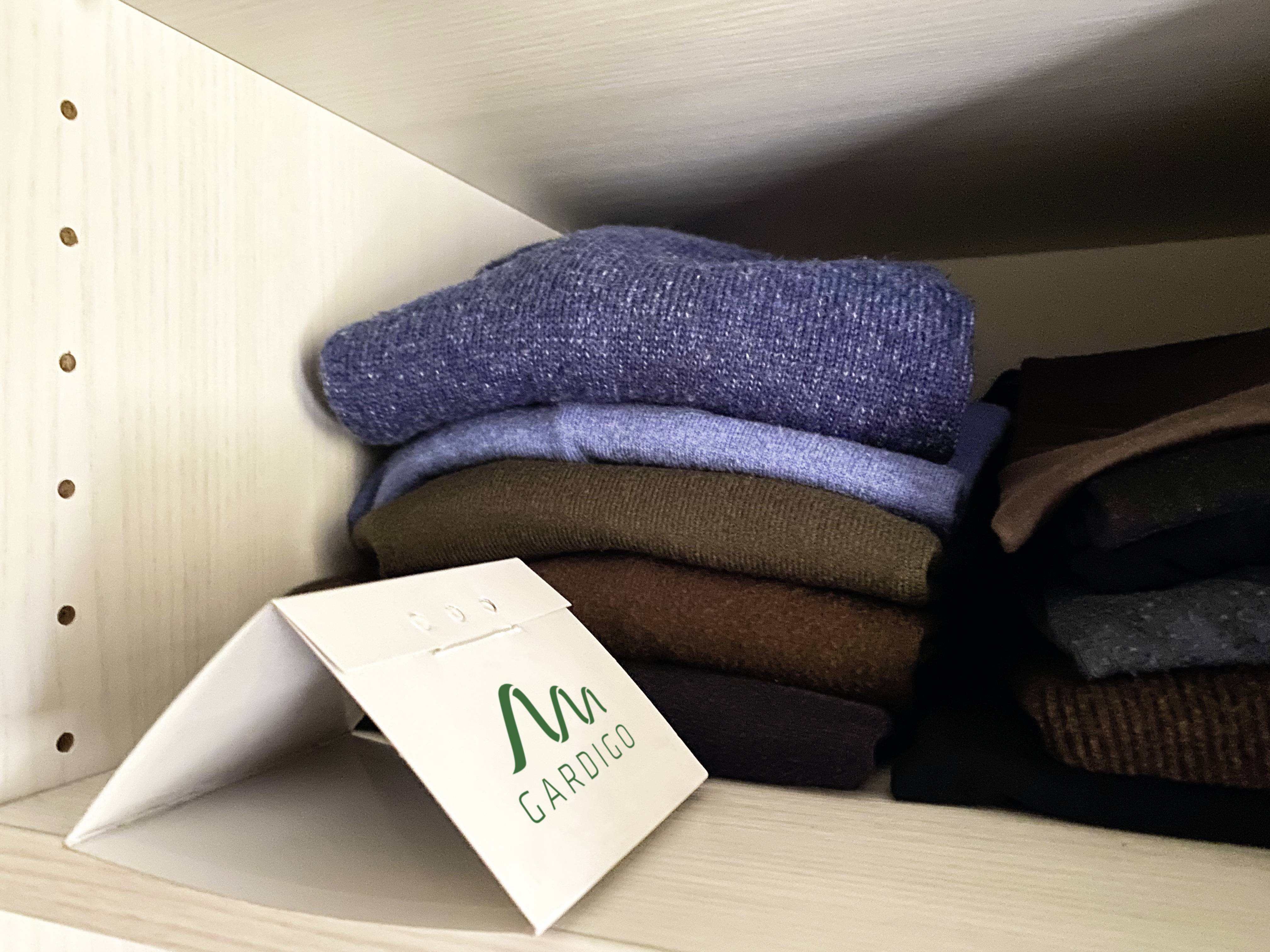 Ein weißes Regal mit mehreren gefalteten Pullovern in verschiedenen Farben, darunter Blau, Braun und Grau. Ein Papieraufsteller mit dem Logo 'Gardigo' steht schräg davor.