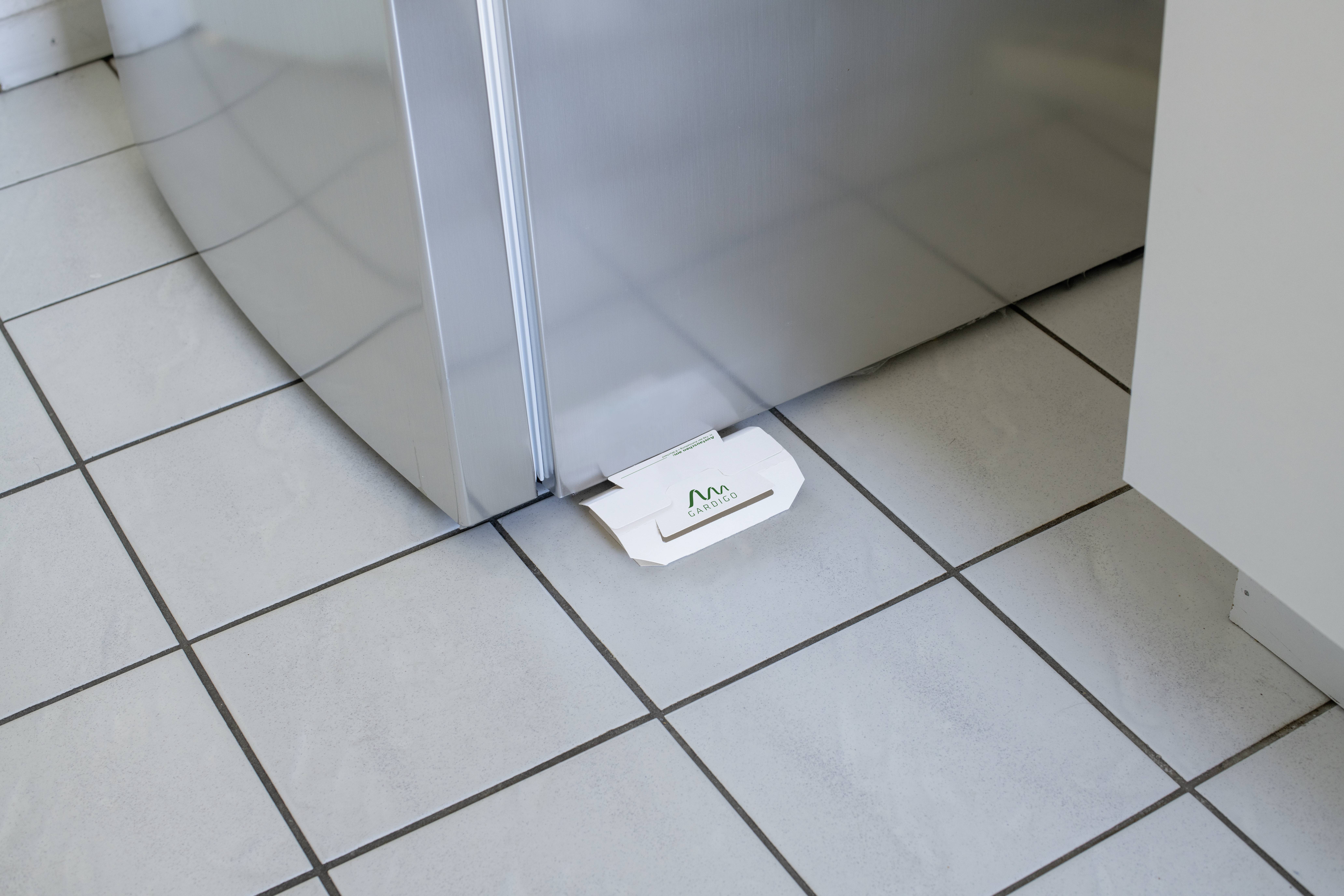 Eine weiße Mausefalle ist auf einem gefliesten Küchenboden vor einem silbernen Kühlschrank platziert.