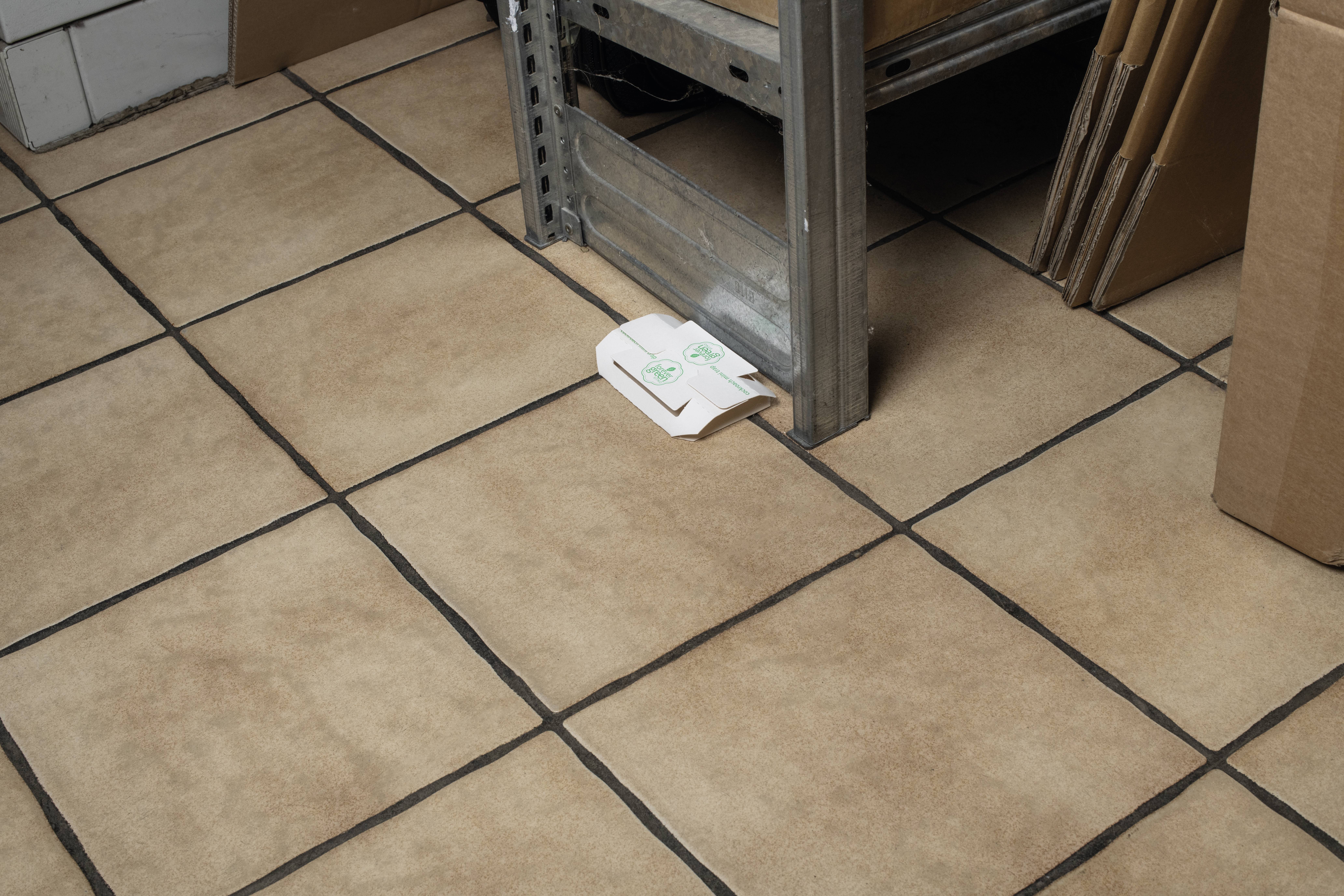 Ein Pappbecherhalter klemmt unter einem Regalbein auf einem gefliesten Boden fest. Daneben stehen Pappkartons.