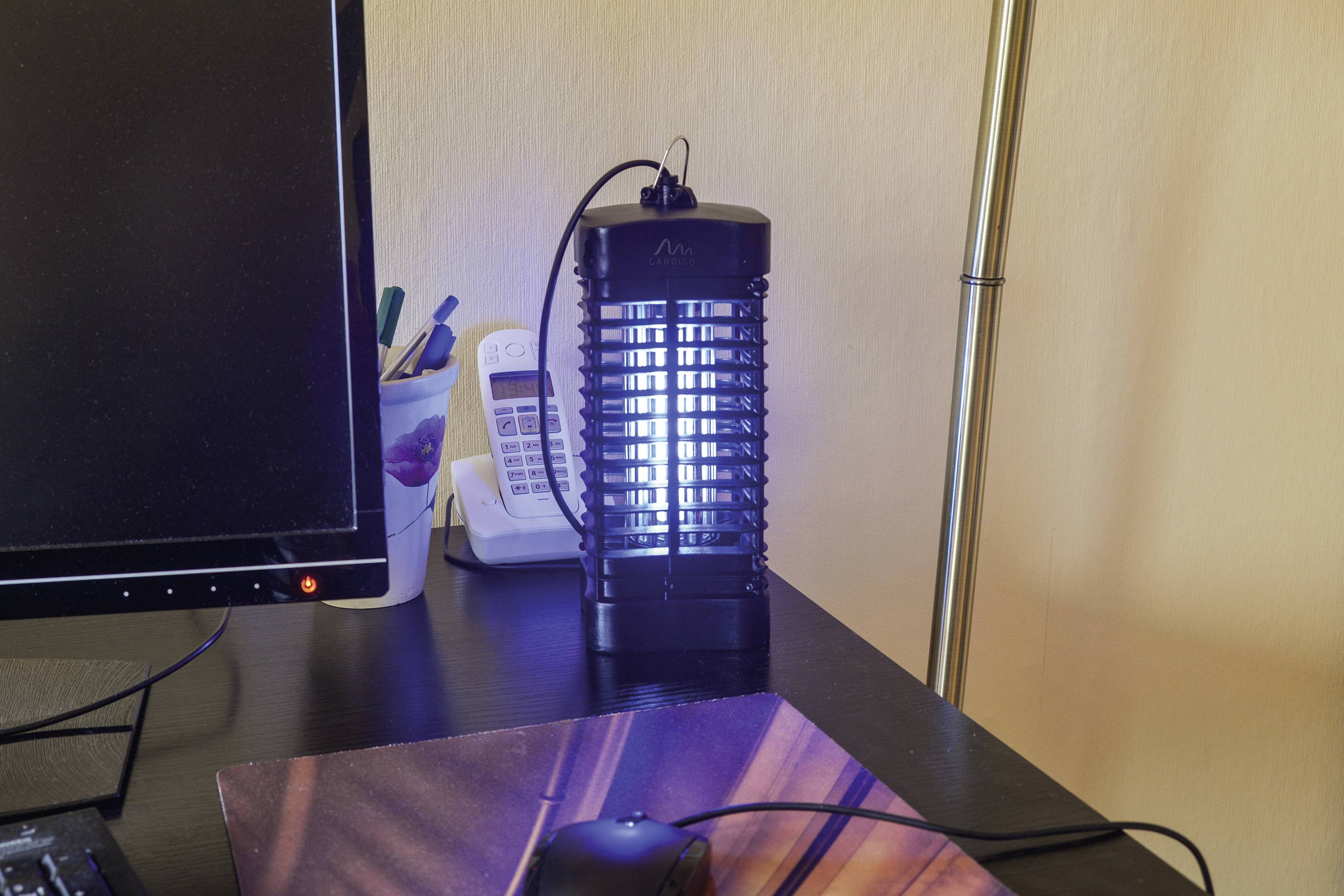 Ein elektrischer Insektenvernichter steht auf einem Tisch neben einem Telefon und einem Computerbildschirm, leuchtet blau.