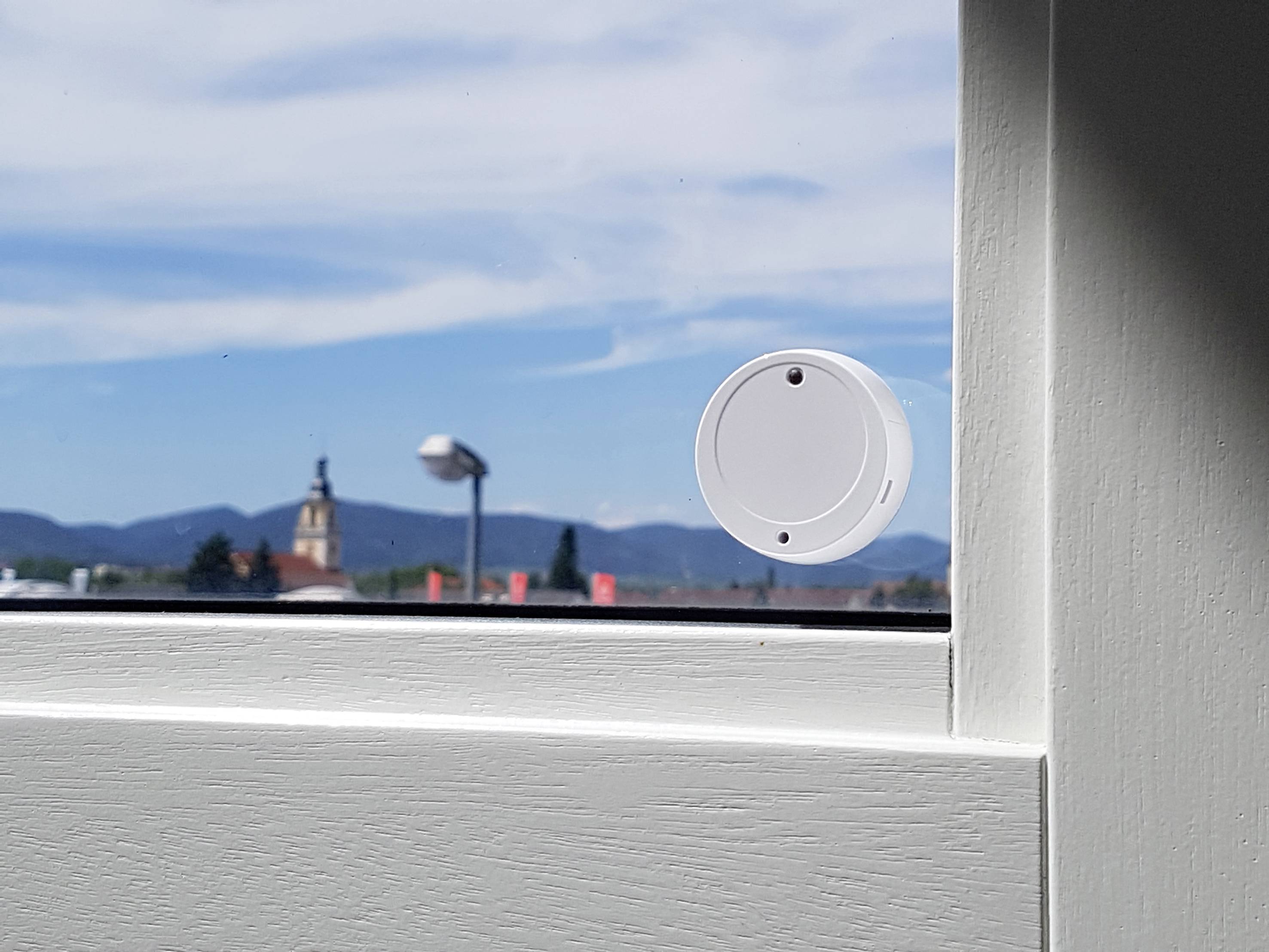 Ein weißer runder Sensor ist an einem Fenster angebracht. Im Hintergrund eine Kirche und Berge bei sonnigem Wetter.