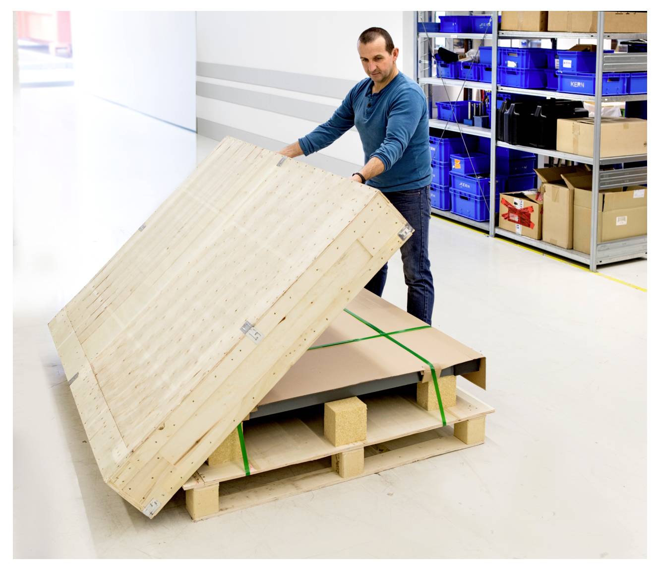Ein Mann hebt einen Holzkistendeckel an, um eine paletierte Sendung in einem Lagerhaus zu inspizieren, mit blauen Lagerkisten auf Regalen im Hintergrund.