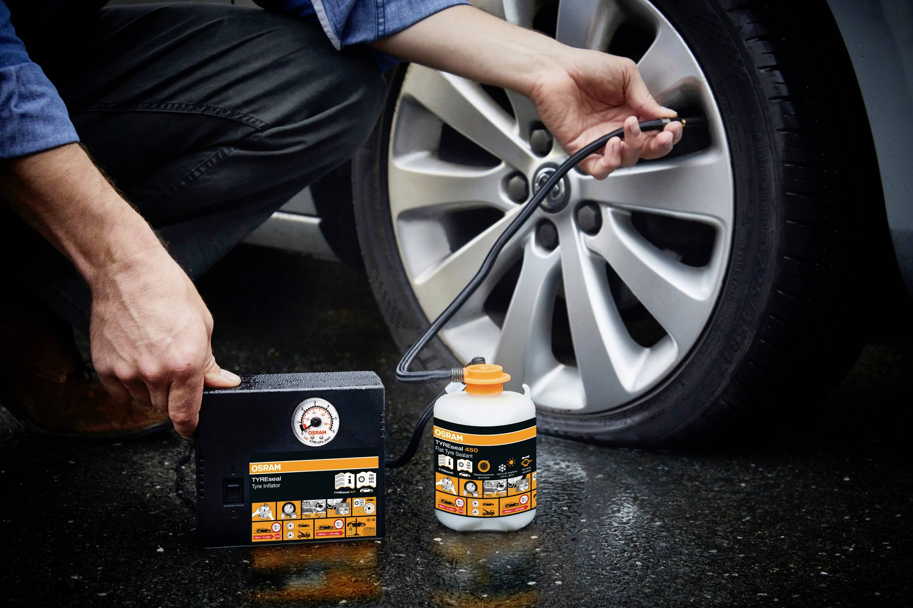 Eine Person repariert einen Autoreifen mit einem Pannenset, bestehend aus einem Kompressor und einer Flüssigkeit. Reifen besitzt 5 Speichen.
