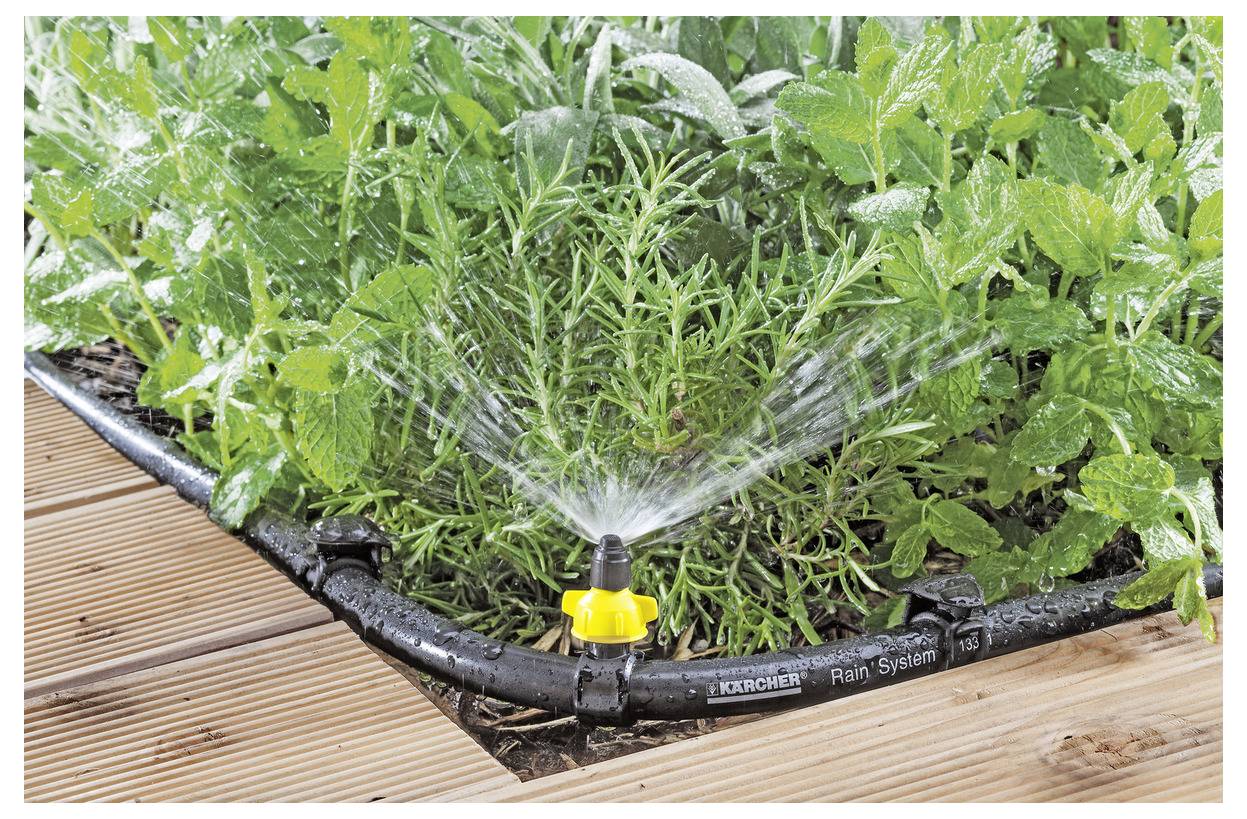Ein Gartenbewässerungssystem, das grüne Pflanzen neben einer Holzterrasse bewässert und ein effizientes Bewässerungssystem demonstriert.