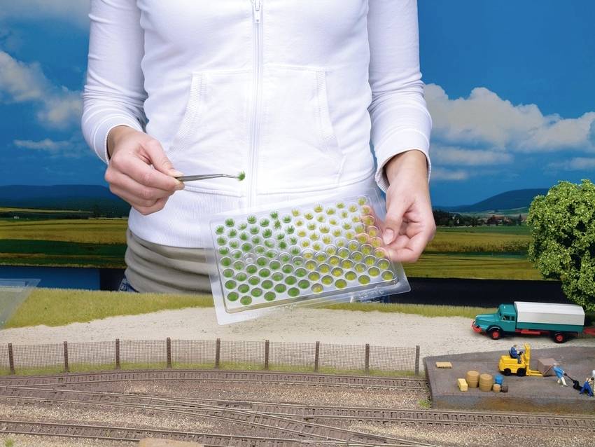 Eine Person platziert mit einer Pinzette grüne Landschaftselemente auf ein Miniaturmodell mit Schienen und einem Lastwagen im Hintergrund.