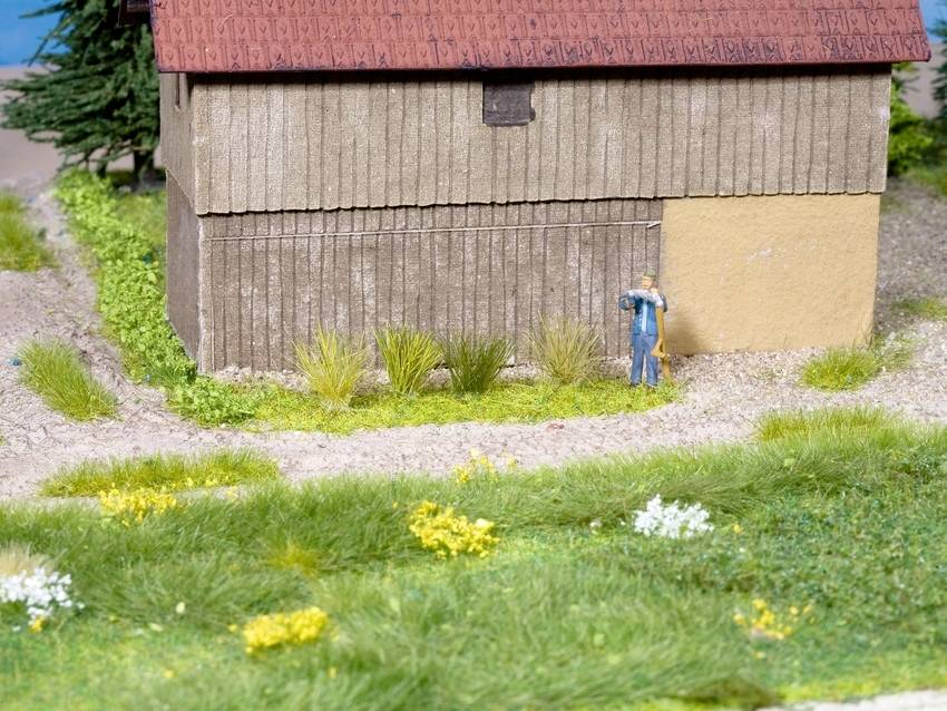 Altes Bauernhaus mit rotem Ziegeldach, umgeben von grüner Wiese und gelben Blumen. Eine kleine Figur lehnt sich an die Hauswand.