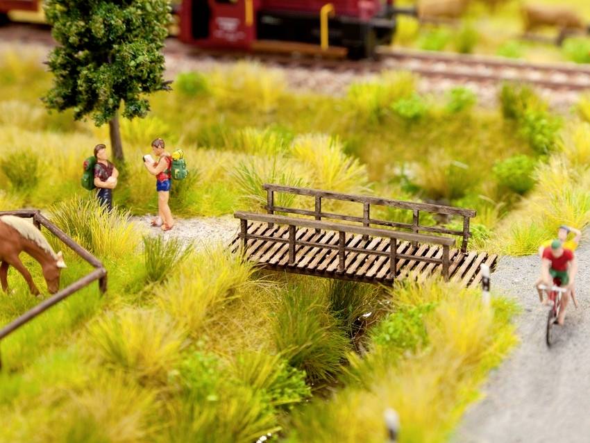 Eine Miniaturlandschaft zeigt Reisende auf einem Weg nahe einer kleinen Brücke, mit einem Pferd im Gehege und einem Fahrradfahrer.