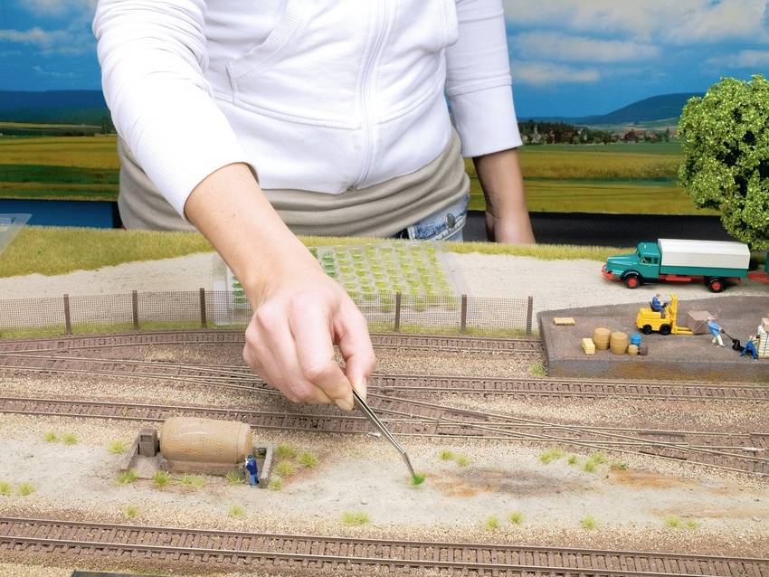 Eine Person gestaltet ein Modellbahn-Diorama mit Gleisen und Landschaftselementen. Im Hintergrund sind Figuren und ein Lastwagen zu sehen.