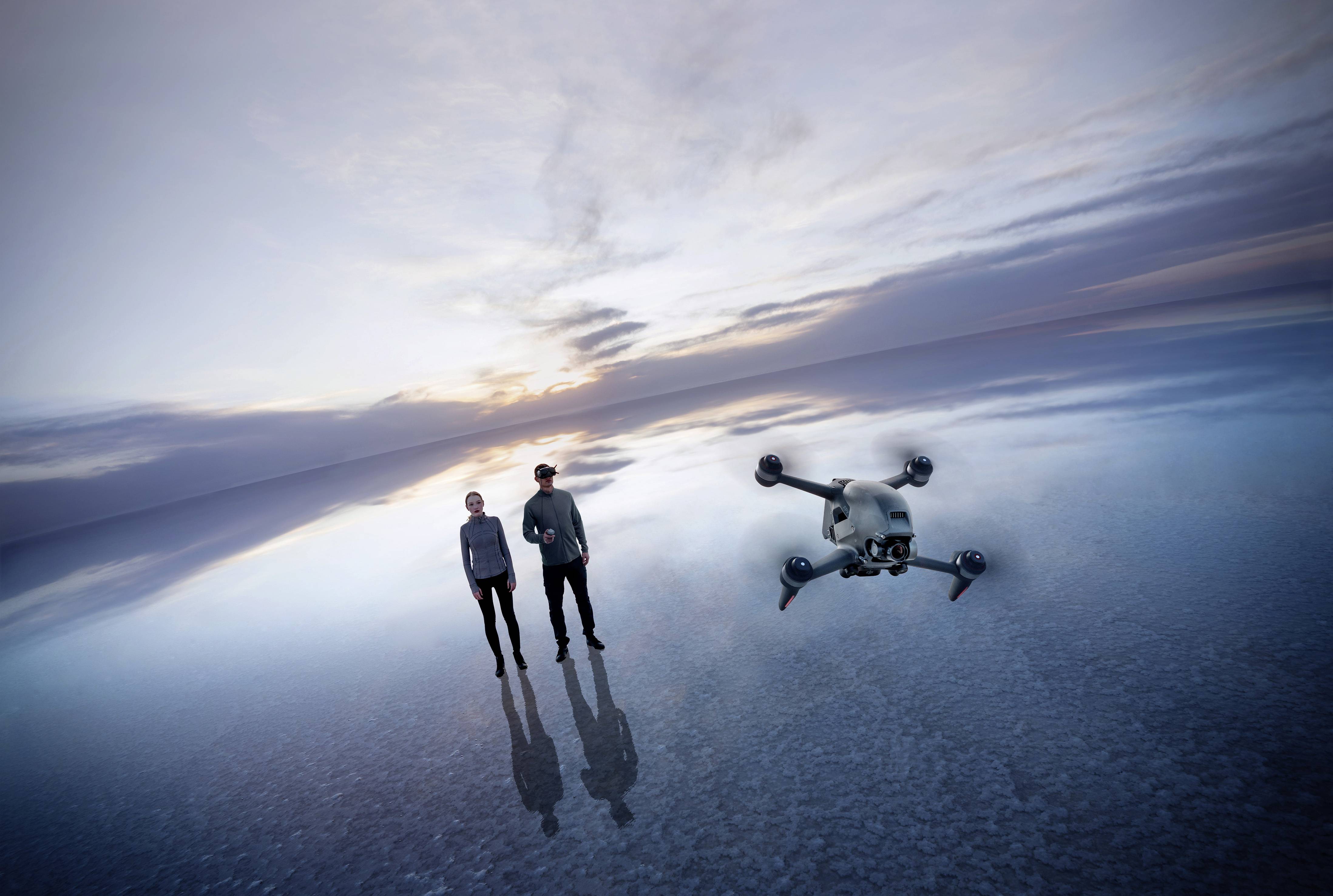 Zwei Personen stehen auf einer spiegelnden Wasseroberfläche bei Sonnenuntergang, während eine Drohne in der Luft schwebt.