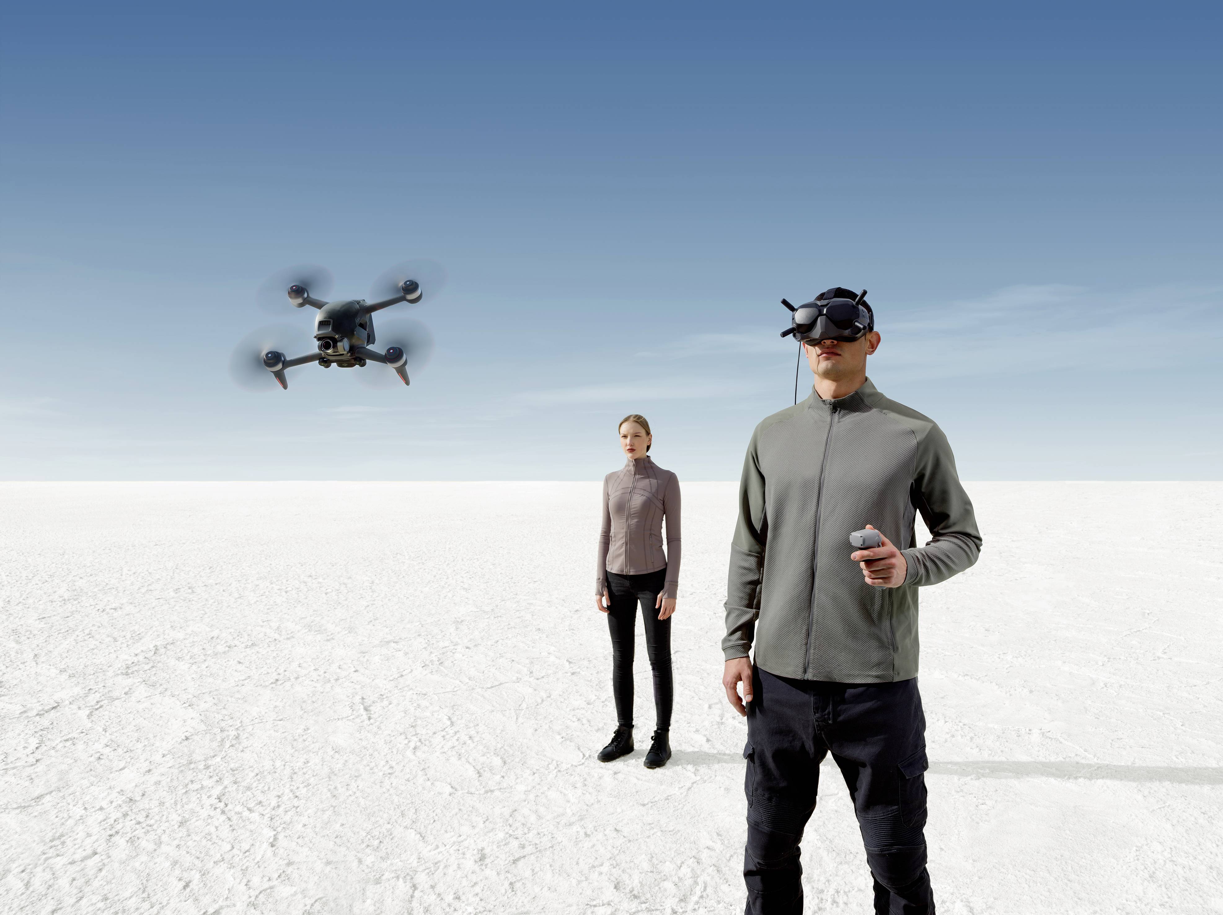 Ein Mann mit VR-Brille fliegt eine Drohne in einer weiten weißen Landschaft, daneben steht eine Frau. Der Himmel ist klar und blau.
