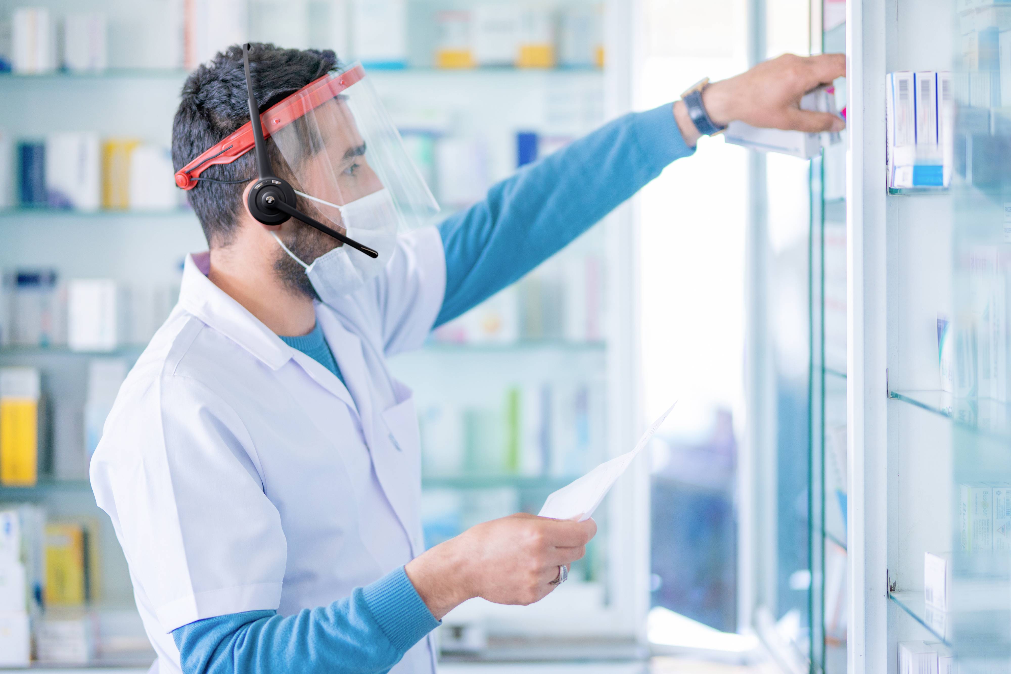 Ein Apotheker trägt Gesichtsschutz und ein Headset, während er Medikamente aus einem Regal nimmt, und überprüft dabei eine Liste.