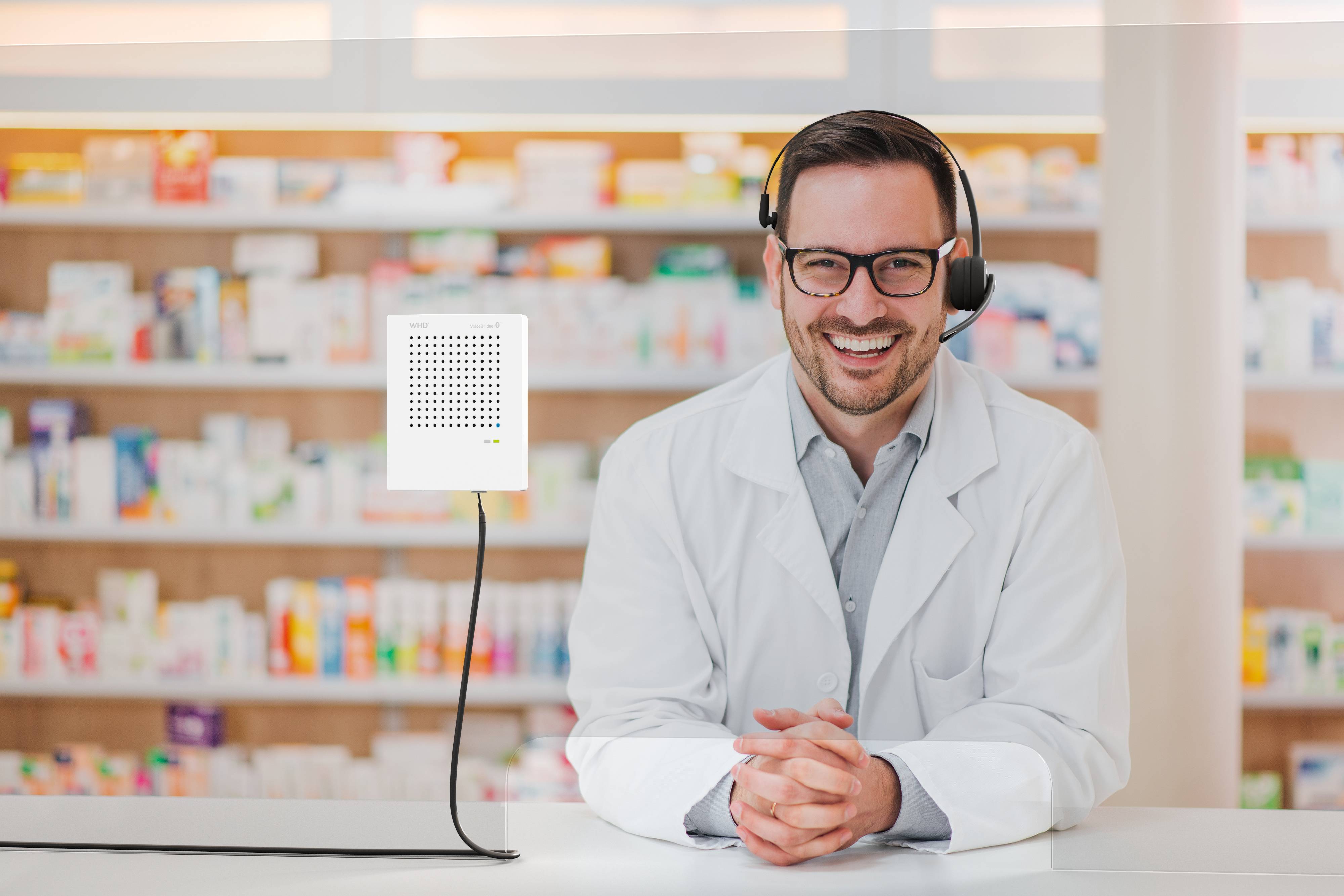 Ein lächelnder Apotheker in weißem Kittel steht an einem Tresen und trägt ein Headset. Im Hintergrund sind Regale mit Medikamentschachteln.