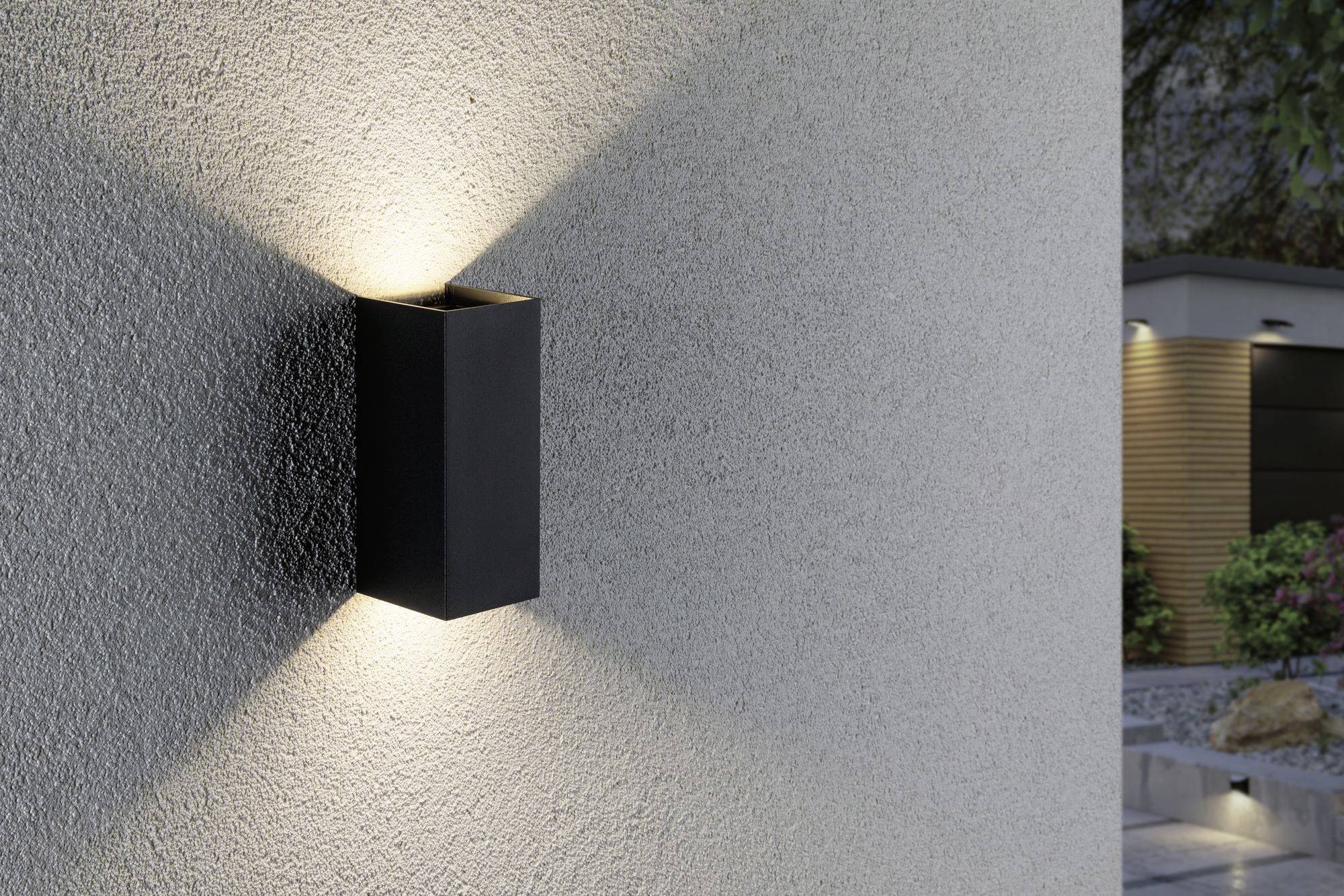 Eine moderne Außenleuchte mit schwarzem Gehäuse strahlt Licht nach oben und unten an einer strukturierten grauen Wand entlang aus.