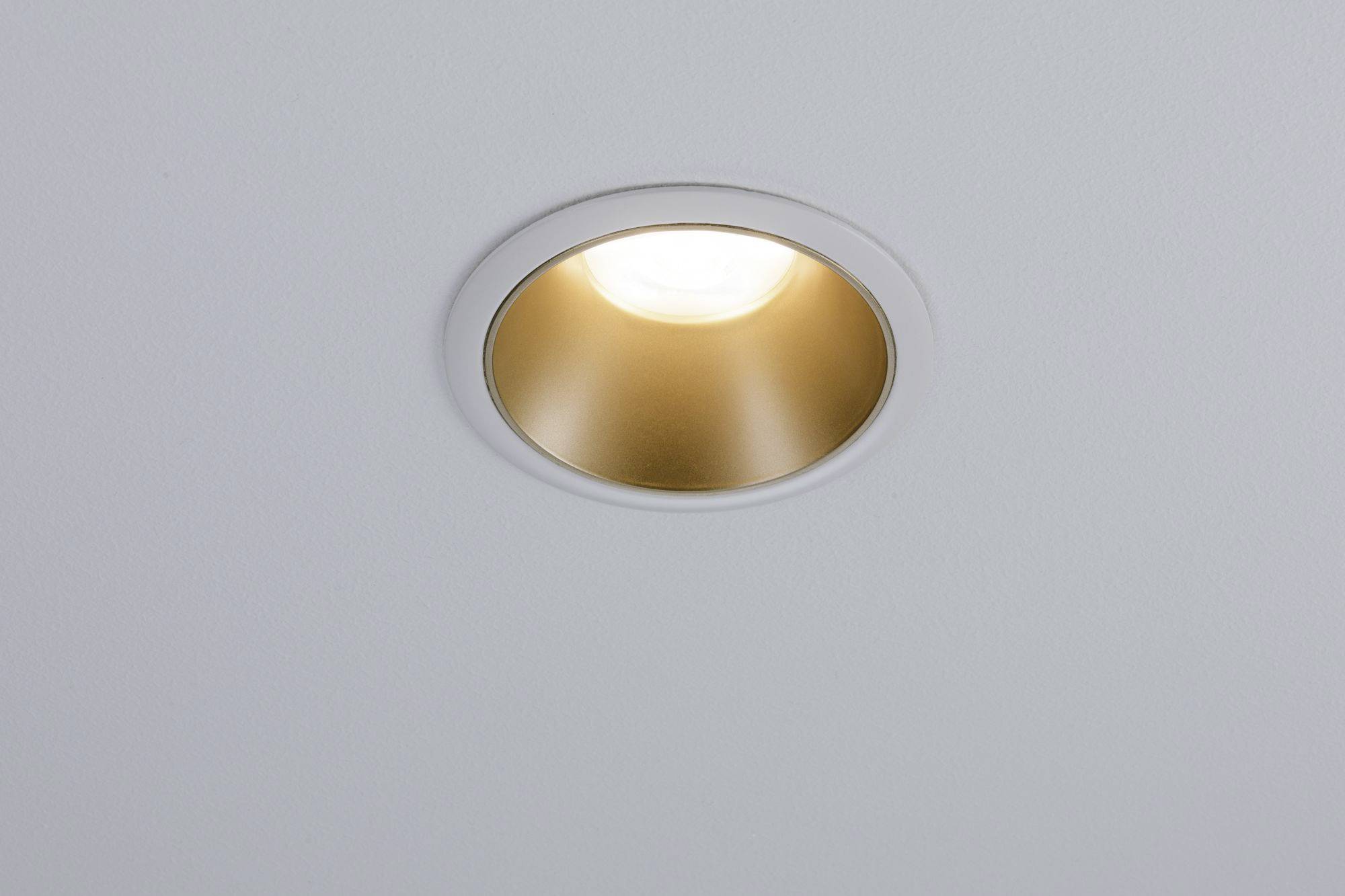 Eine runde, eingelassene Deckenleuchte mit goldener Fassung leuchtet, in einem schlichten Raum installiert.