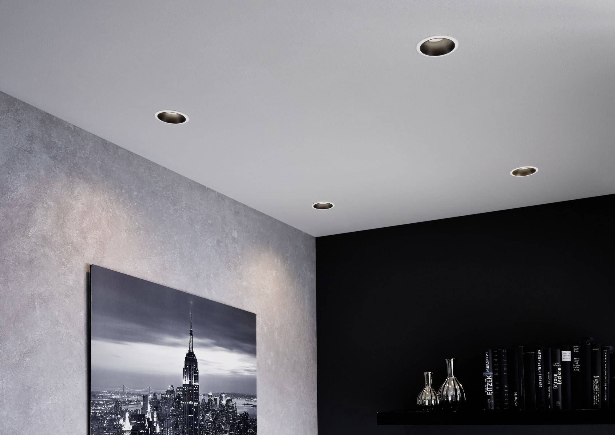Innenraum mit schwarzer Wand und eingebauten Deckenstrahlern. Ein Foto von einer Skyline hängt an der Wand; daneben Regale mit Büchern.