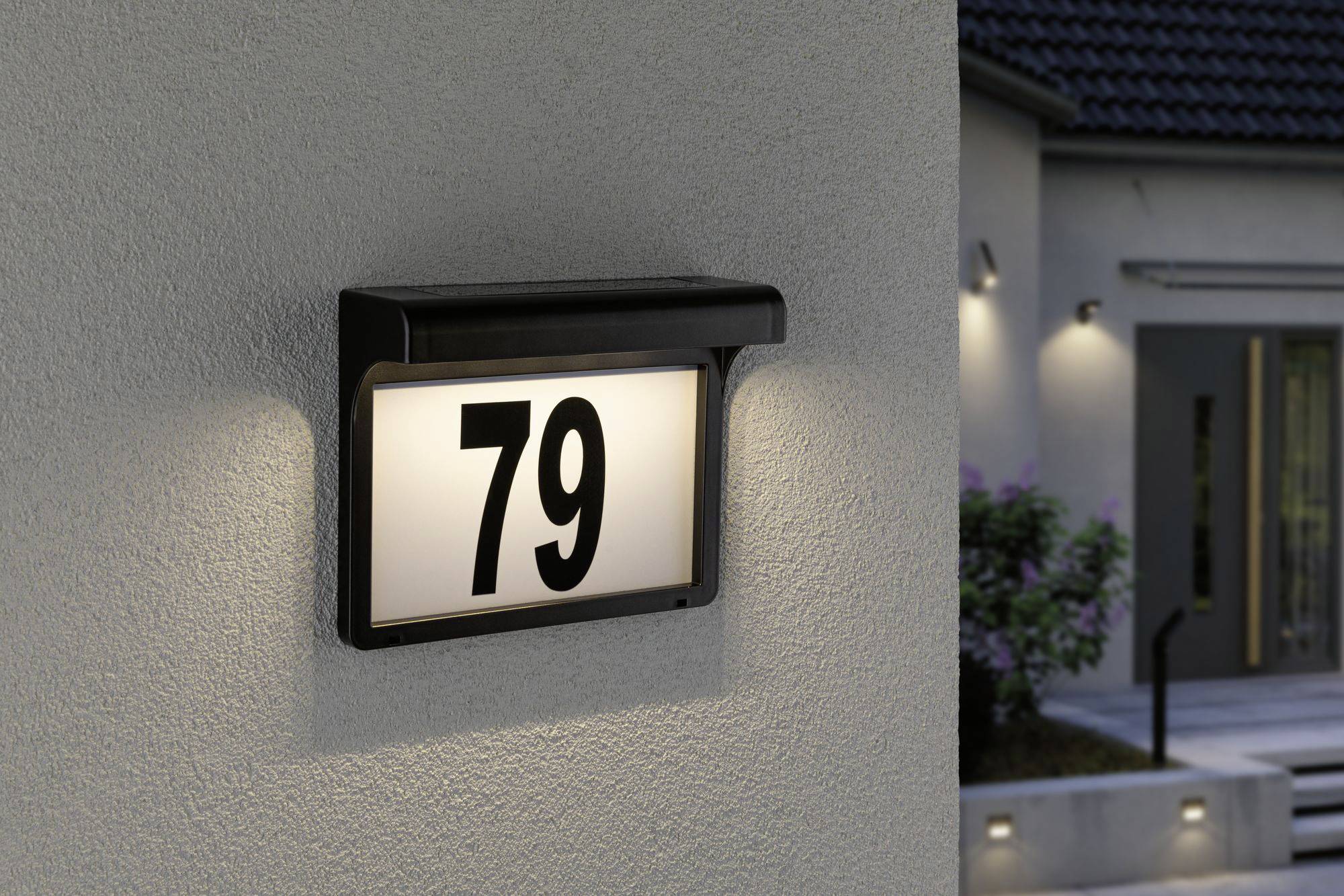 'Beleuchtetes Hausnummernschild 79 an einer hellen Wand; im Hintergrund sind verschwommene Konturen eines Gebäudes sichtbar.'