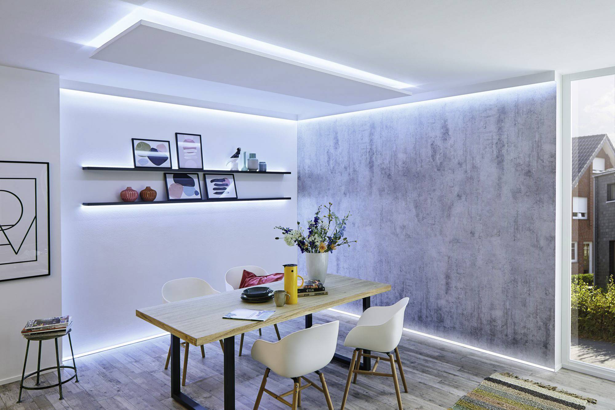 Moderner Essbereich mit weißem Tisch und Stühlen, beleuchtet von LED-Streifen. Regale mit Dekorationen an der Wand, große Fenster.