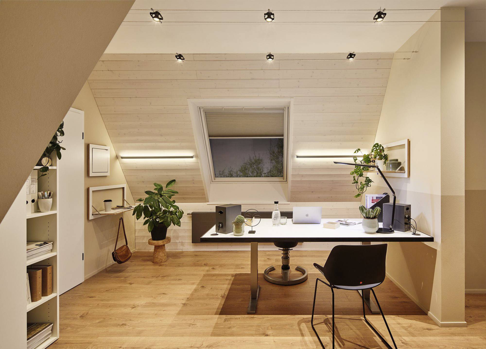 Ein modern eingerichtetes Homeoffice mit einem großen Schreibtisch, Laptop, zwei Lautsprechern, Pflanzen und einem Bücherregal.