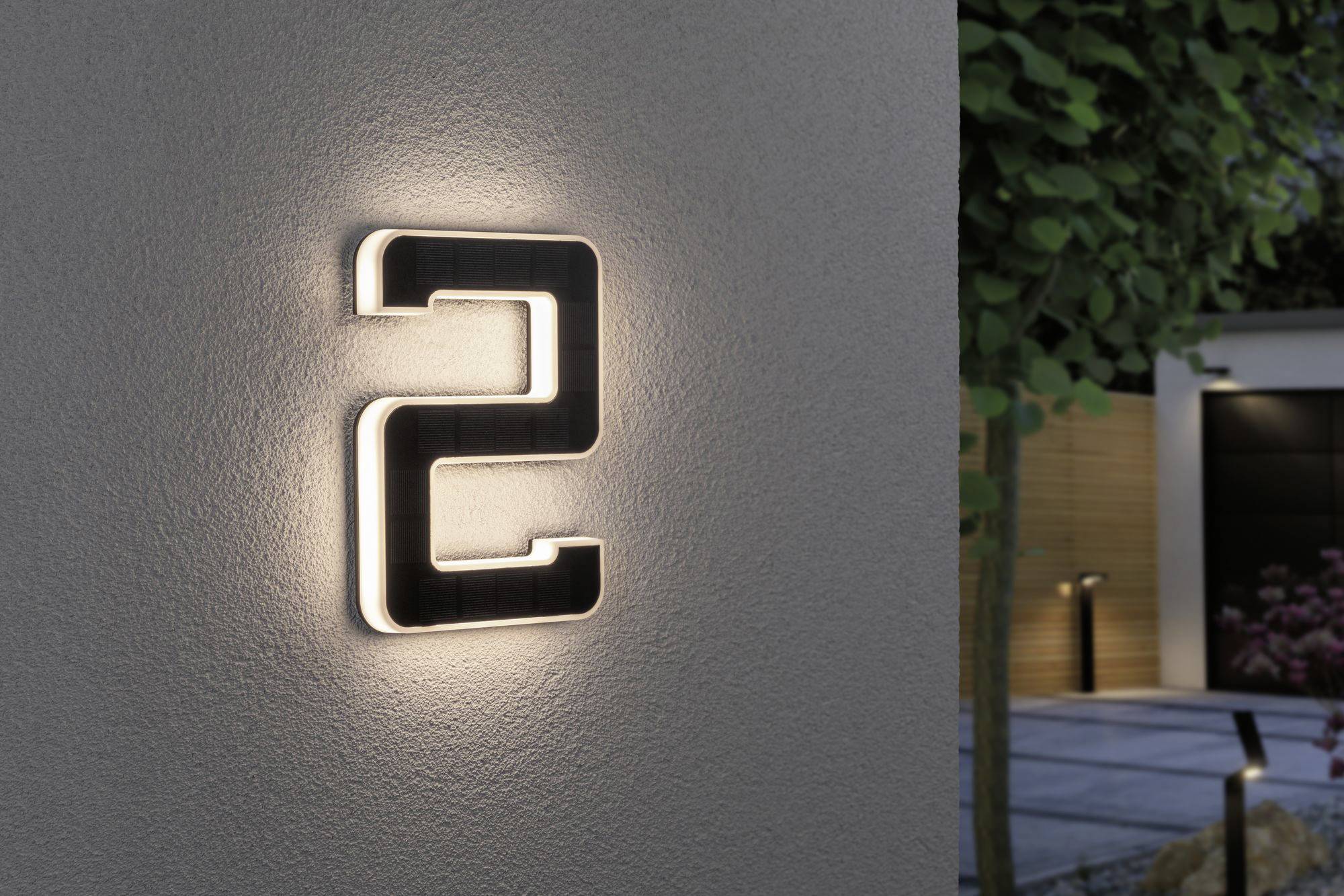 Leuchtende Hausnummer '2' an einer grauen Wand, umgeben von einem modernen Garten mit Bäumen und gepflastertem Gehweg.