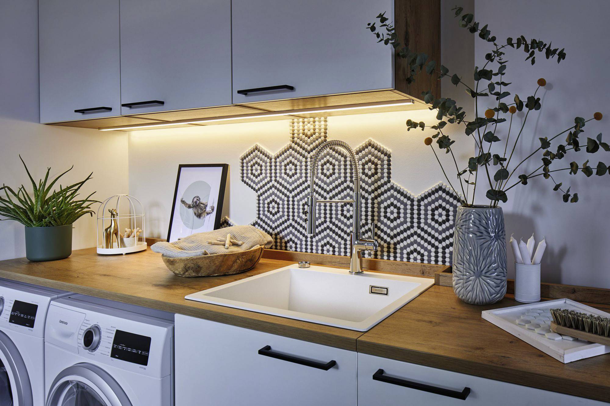 Waschküche mit Arbeitsplatte aus Holz, Spüle in der Mitte, dekorativem Fliesenmuster an der Wand, und Pflanzen-Dekorationen.